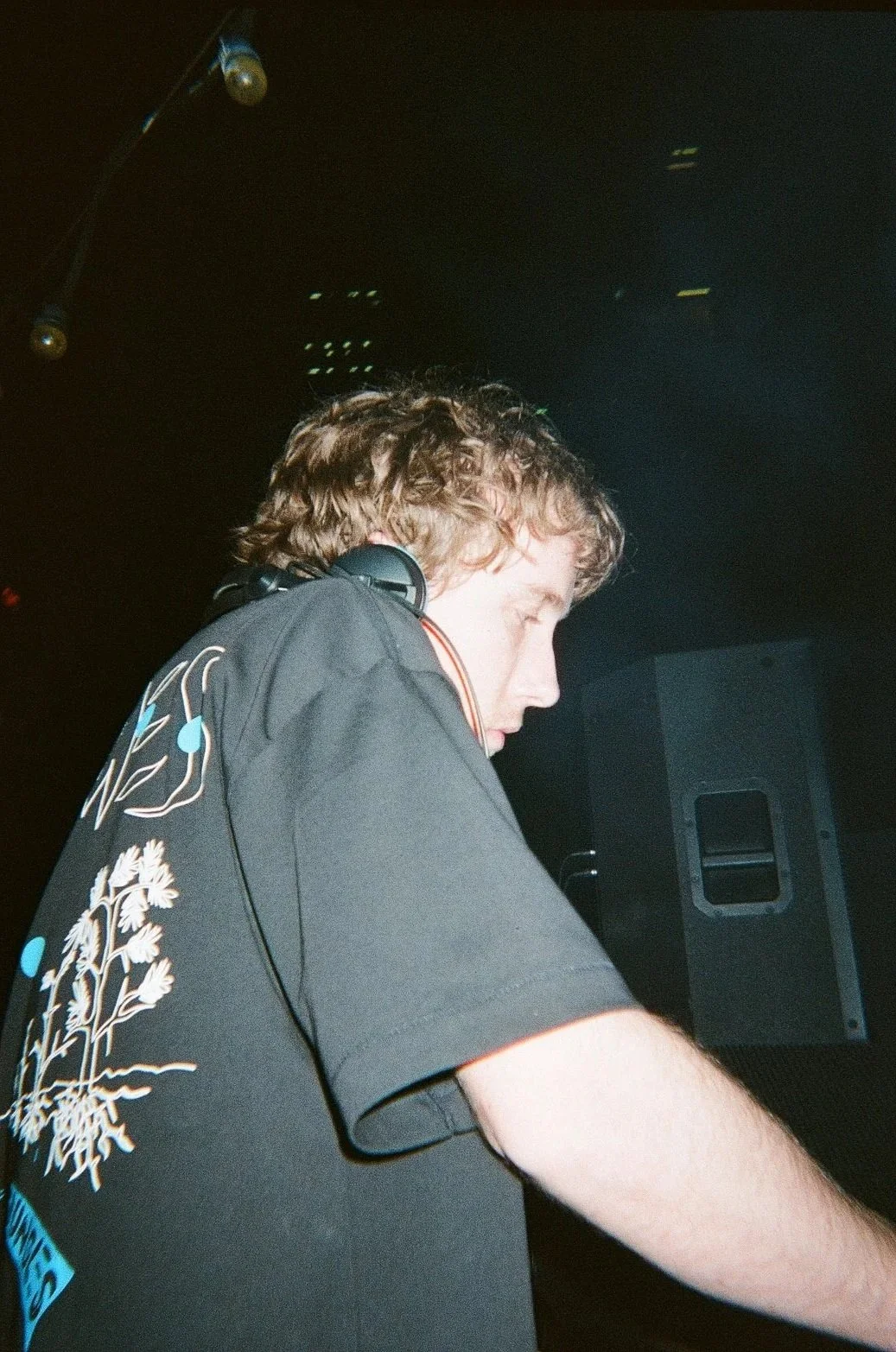 A young male DJ or music producer with curly hair, wearing a black shirt with decorative print on the shoulder, is focused on his equipment in a dark setting.