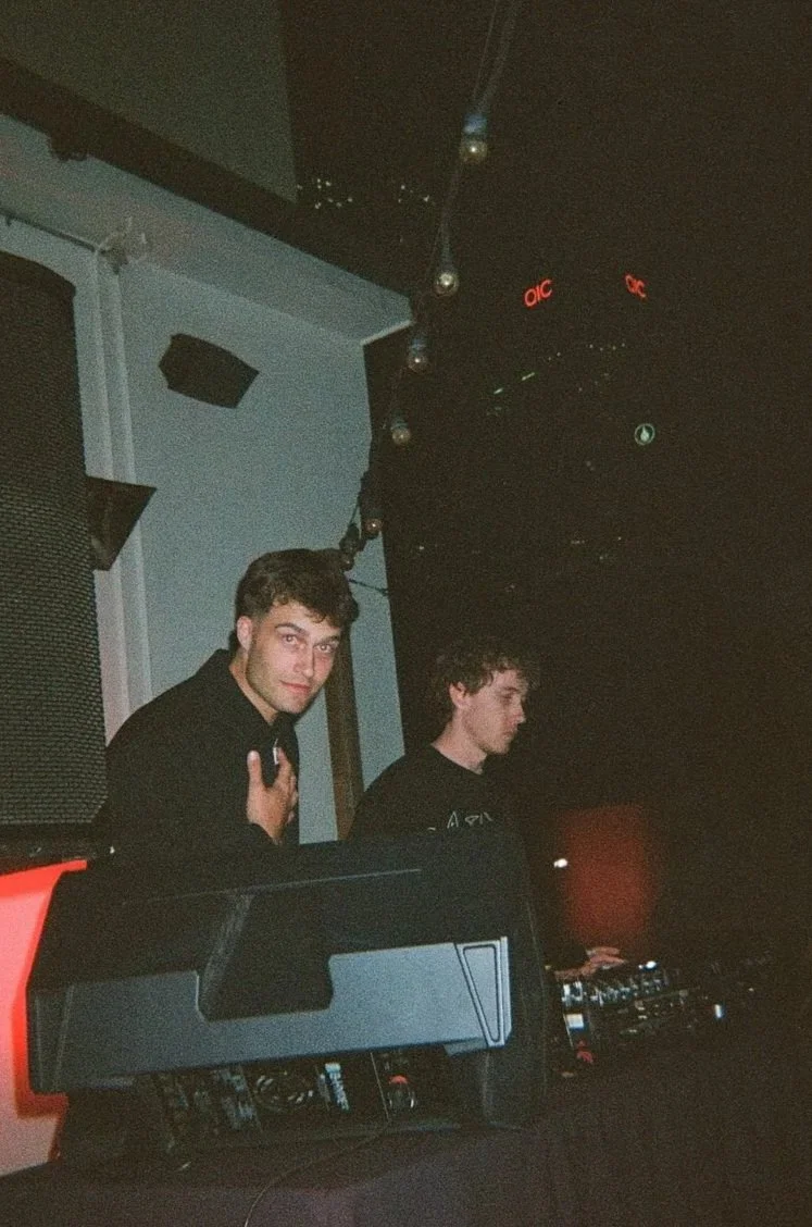 Two young men are DJing in a dark club or venue with music equipment and black walls, one is looking at the camera with a hand on his chest, the other is focused on the DJ equipment.