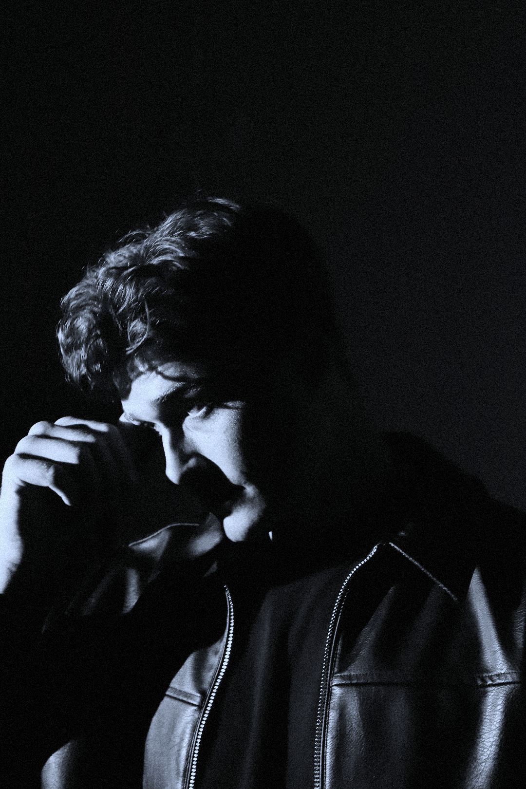 A black-and-white photo of a young man with curly hair, wearing a leather jacket, with his hand touching his forehead, looking downward in a contemplative pose.