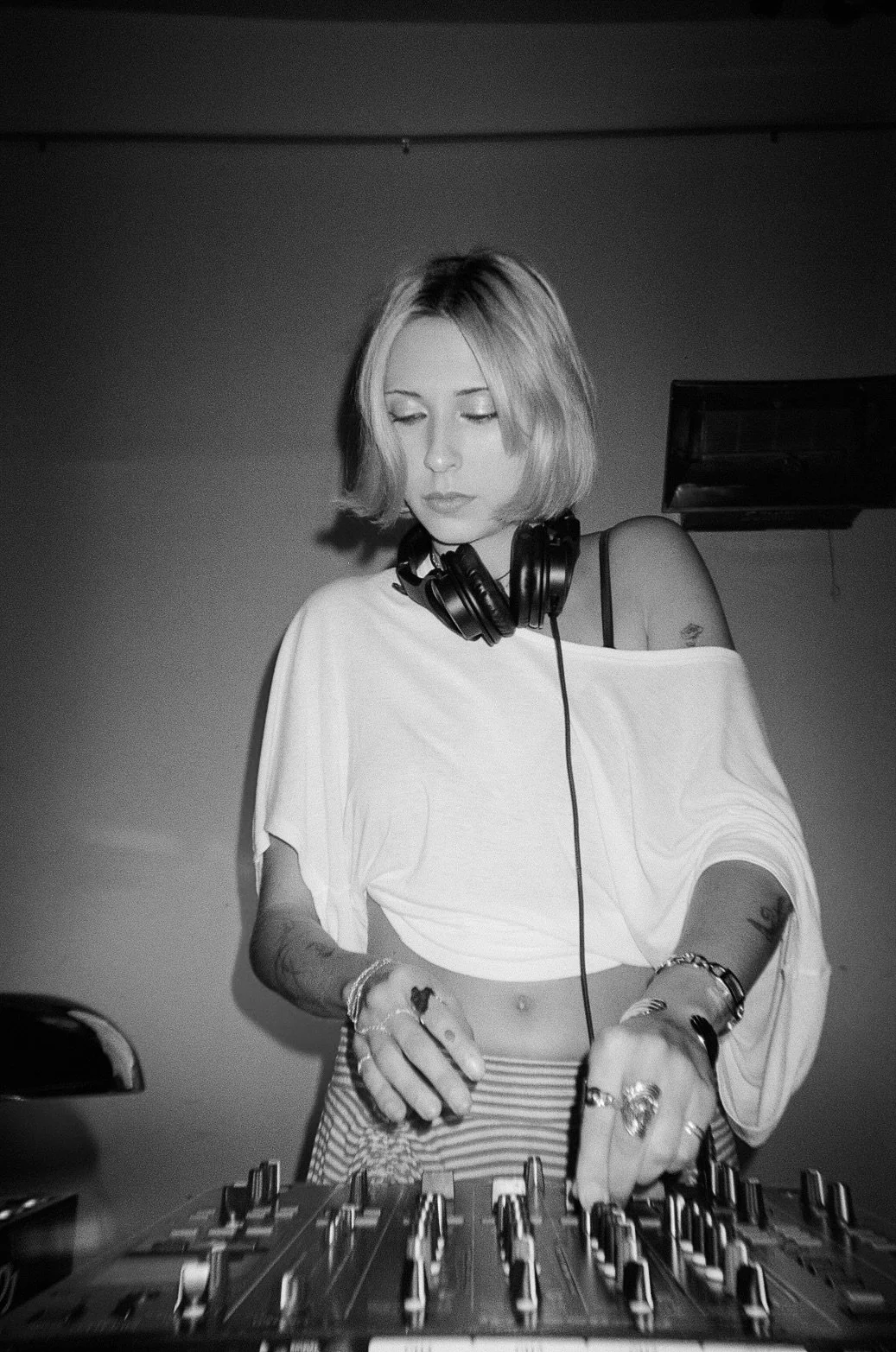 A young woman with short blond hair wearing headphones around her neck, a loose off-shoulder t-shirt, and a striped skirt, operating a DJ mixer.