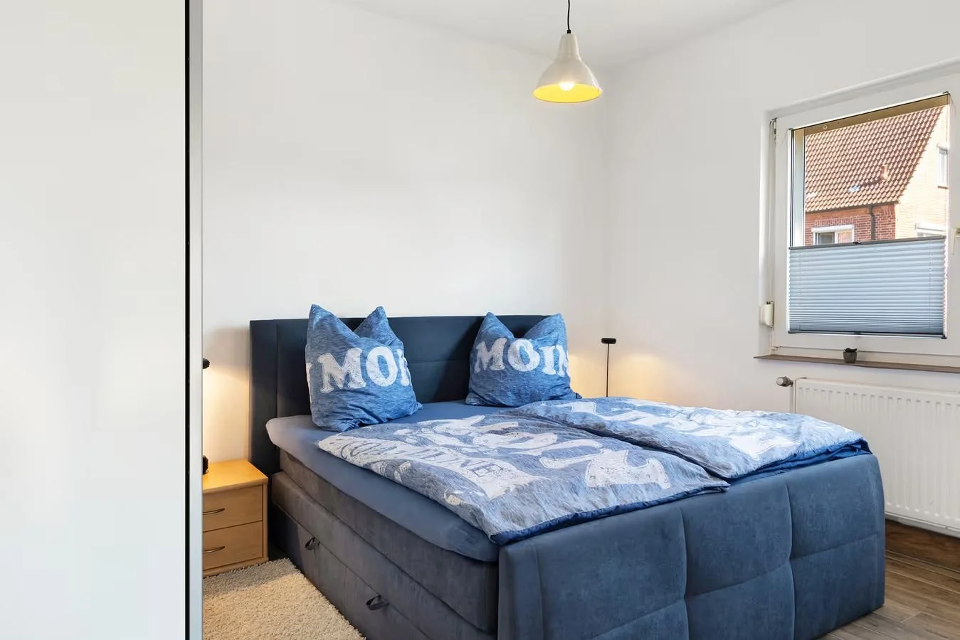 Ein Schlafzimmer mit einem blauen Bett, blauen Kissen mit weißen Buchstaben, einem kleinen Nachttisch, einer Lampe und einem Fenster mit Blick auf ein Haus.