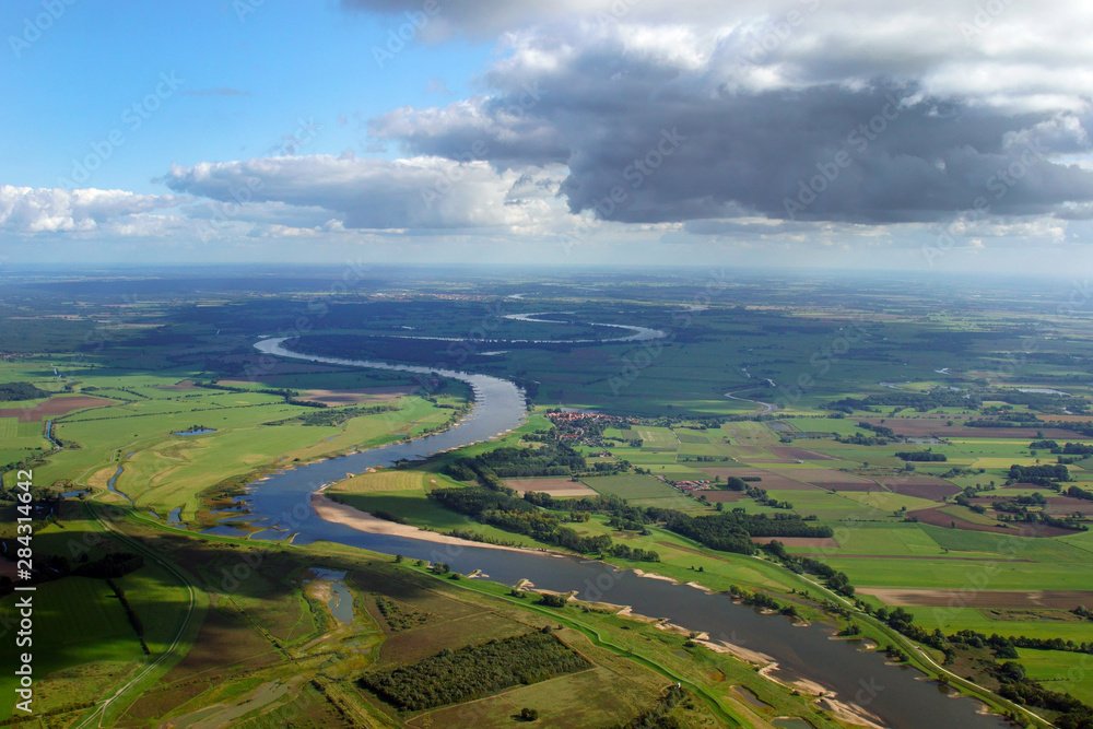 Luftaufnahme eines Flusses, der sich durch eine grüne Landschaft windet, mit Feldern und kleinen Siedlungen, unter einem bewölkten Himmel.