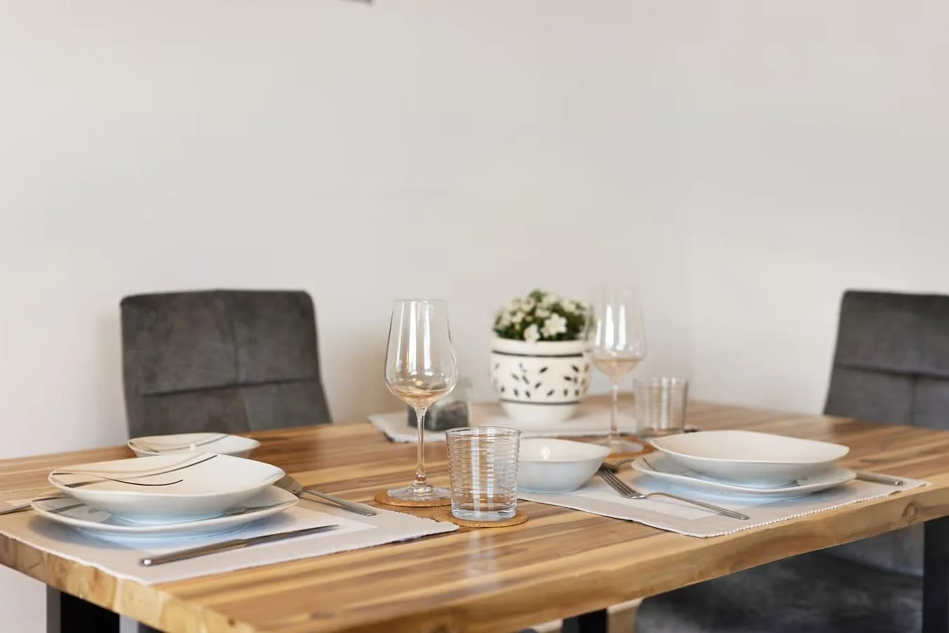 Ein Esstisch mit weißen Tellern, Gläsern und Besteck, eine weiße Blumenvase mit weißen Blumen, Platzsets und Stühlen auf beiden Seiten, schlichtes, modernes Dekor.