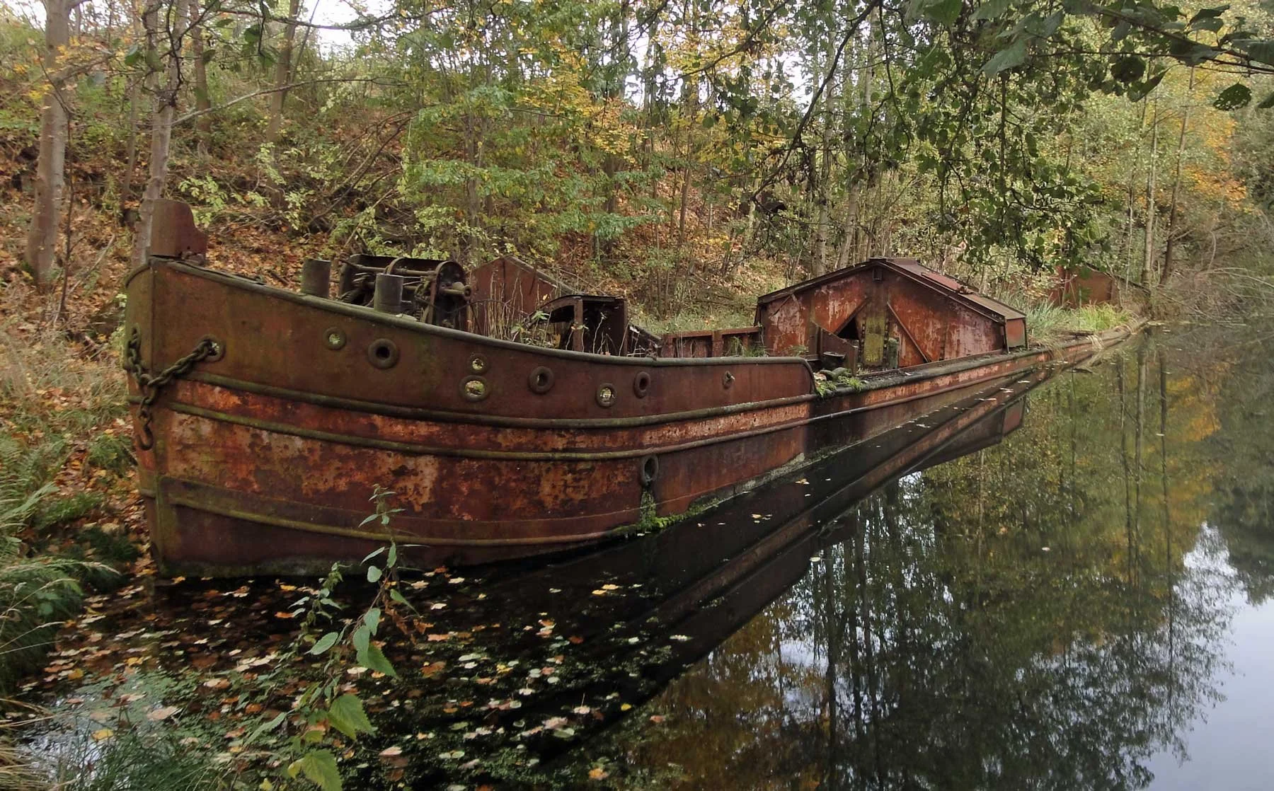 Ein verrostetes, altes Boot liegt im Wasser, umgeben von Bäumen mit Herbstlaub. Das Boot ist still und spiegelt sich im Wasser.