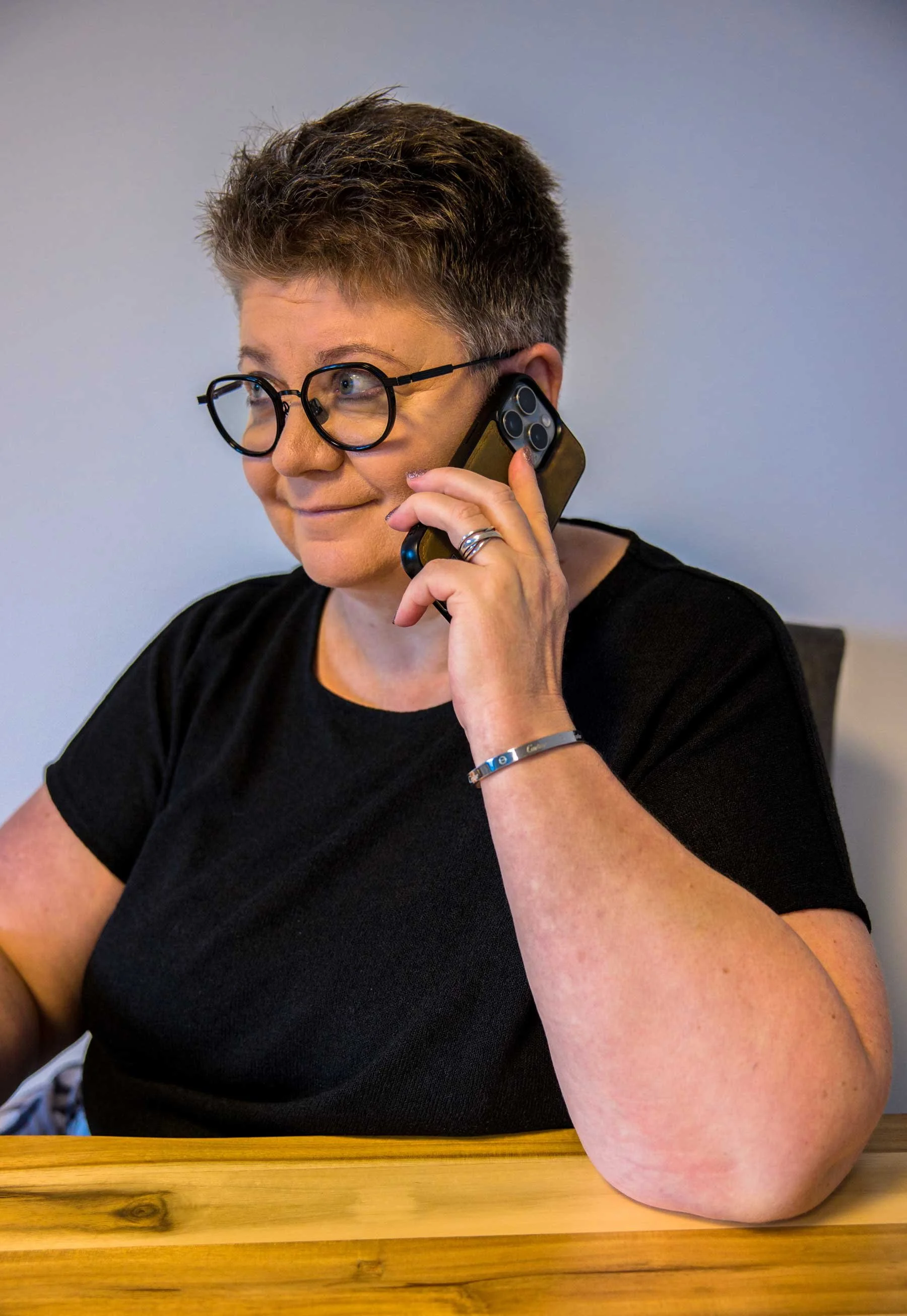 Eine Person mit kurzen, grau-braunen Haaren und schwarzen Brillen spricht am Telefon. Sie trägt ein schwarzes T-Shirt und sitzt an einem Holztisch.