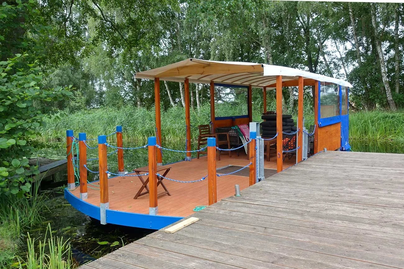 Eine Holzboot mit einem Dach, das an einem Steg an einem Fluss oder Teich liegt, umgeben von grünen Büschen und Bäumen.