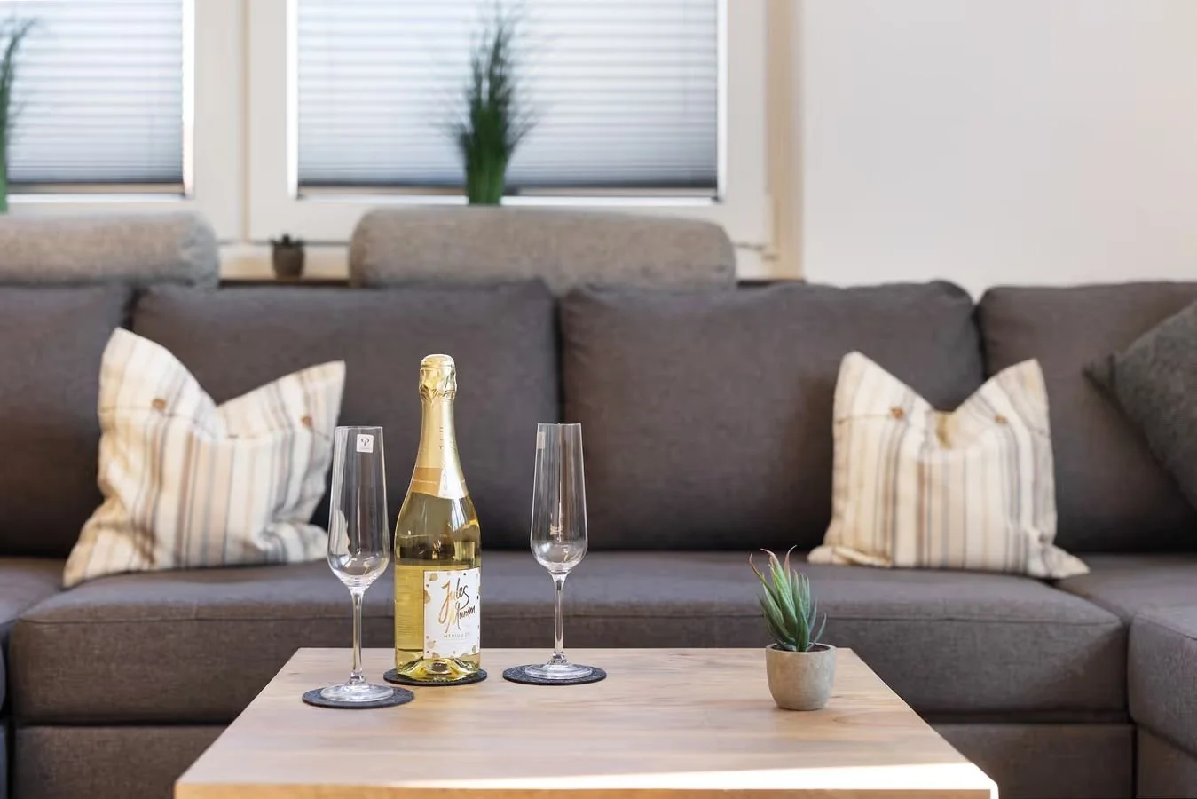 Wohnzimmer mit grauer Couch, zwei Streifen-Kissen, einem kleinen Holztisch mit Sektflasche, zwei Sektgläsern und einer kleinen Topfpflanze, Hintergrund mit Fenstern und Vorhängen.