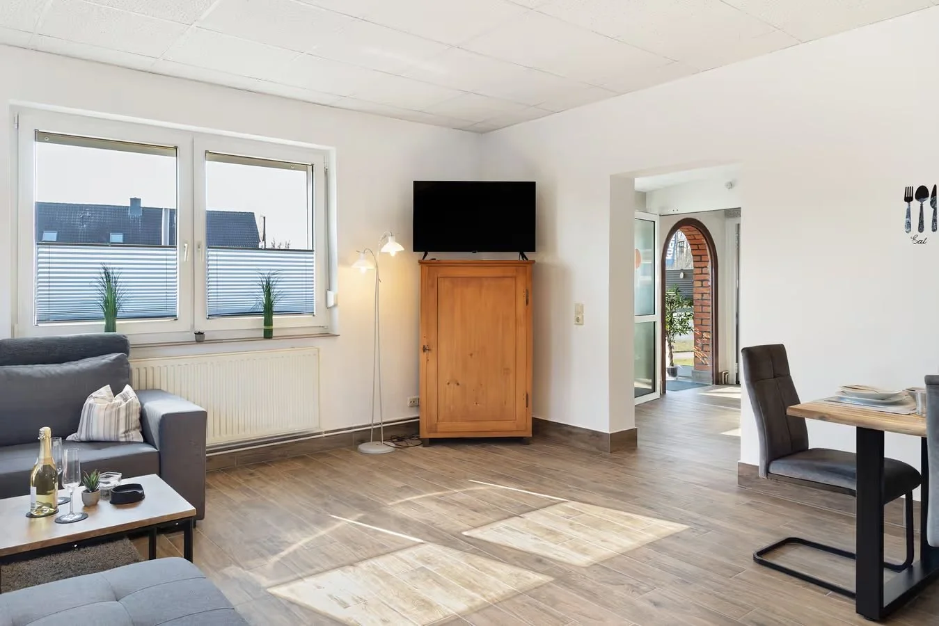 Gemütliches Wohnzimmer mit Holzfußboden, großen Fenstern mit Rollläden, einer grauen Couch, einem kleinen Tisch mit Getränken, einem Fernseher auf einem Holzkasten, Stehlampe, Essbereich mit Stuhl und Tisch, Eingangstür im Hintergrund.