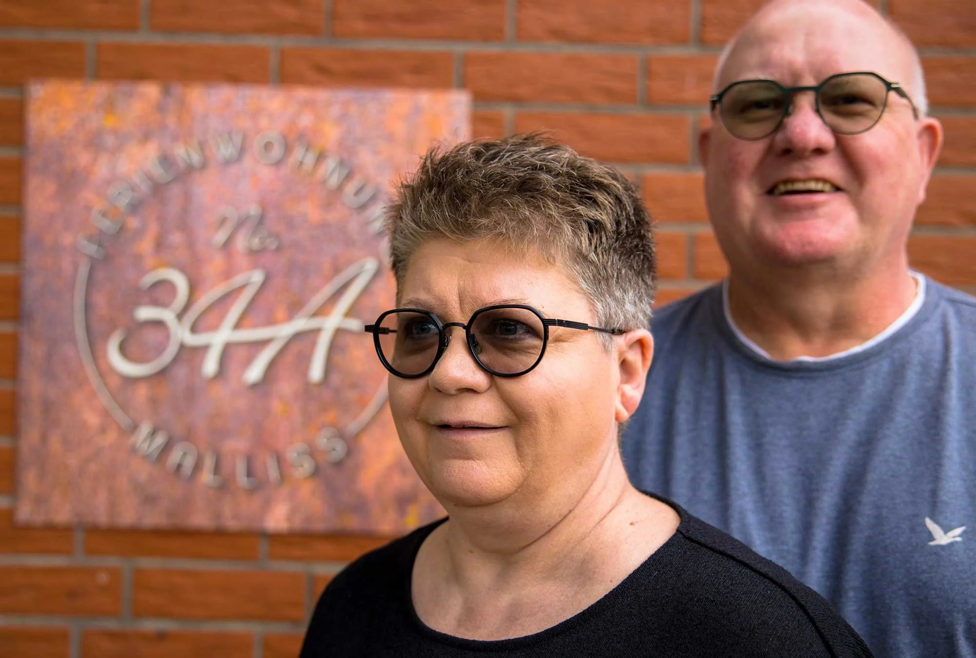 Zwei Menschen, eine Frau im Vordergrund mit kurzen grauen Haaren und schwarzen Brillen, ein Mann im Hintergrund mit kurzen Haaren, Brille und grauem T-Shirt, stehen vor einer roten Backsteinmauer. Beide lächeln.