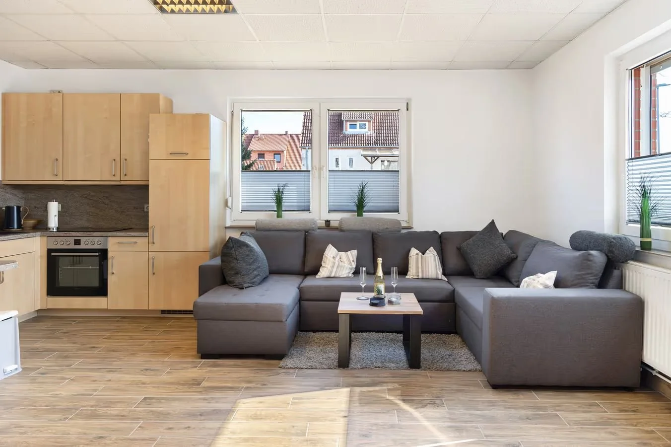 Wohnzimmer mit grauem Sofa, kleinen Kissen, Couchtisch mit Sekt und Gläser, zwei Fenster mit Pflanzen, moderner Einbauküche mit Holzfronten.
