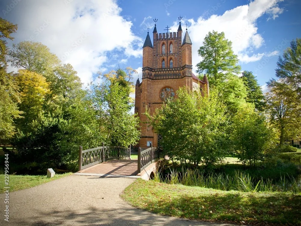 Ein historisches rotes Backsteingebäude mit kurzen Türmen und Fenstern, umgeben von Bäumen, mit einer Brücke im Vordergrund unter einem blauen Himmel mit Wolken.