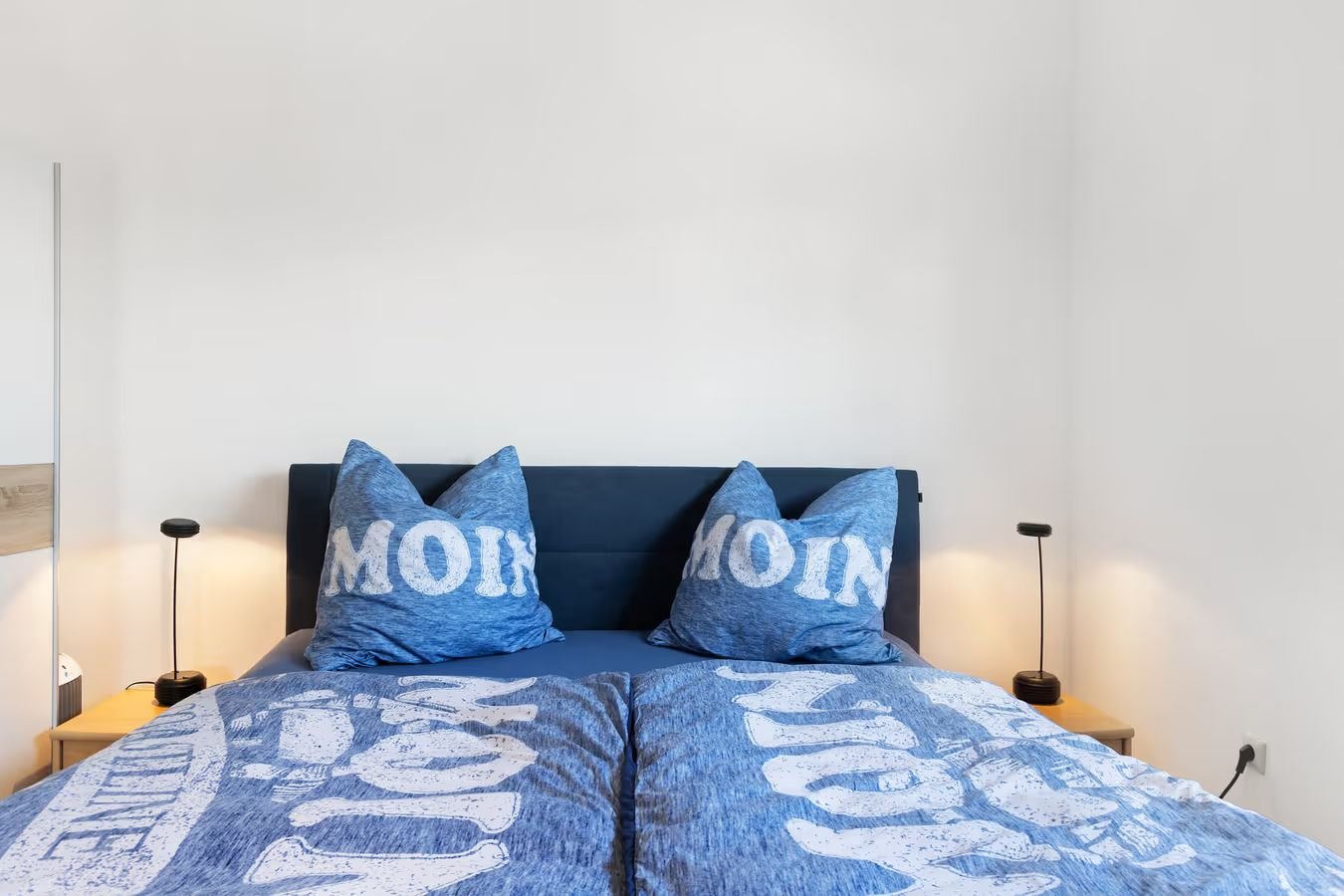 Bett mit blaue Bettwäsche und zwei Kissen mit der Aufschrift "MOM" in einem Schlafzimmer, seitliche Nachttische mit schwarzen Lampen