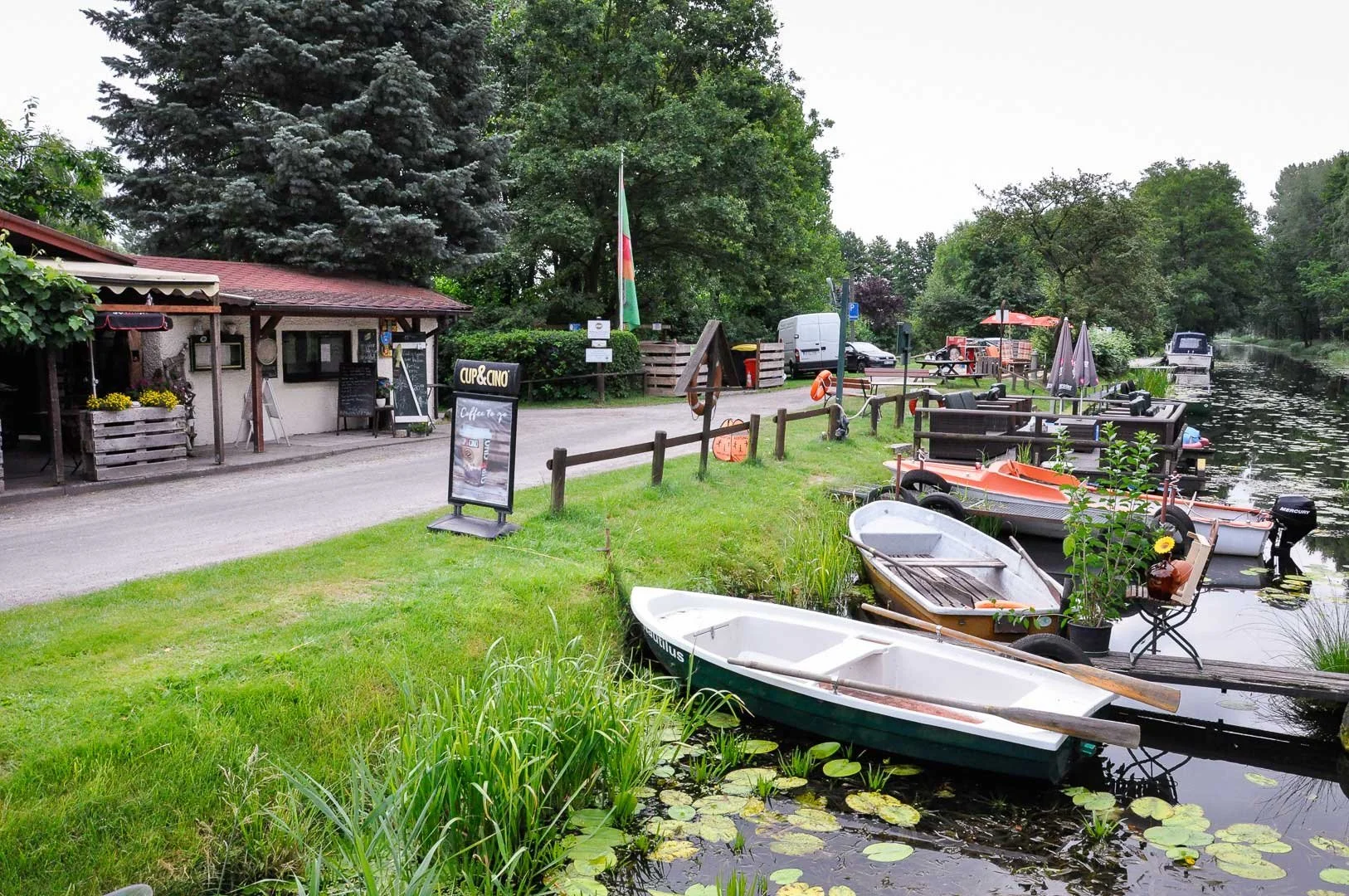 Ein Ufer mit mehreren Boote, die im Wasser liegen, und einem kleinen Restaurant oder Café mit Tischen im Freien, umgeben von Bäumen.