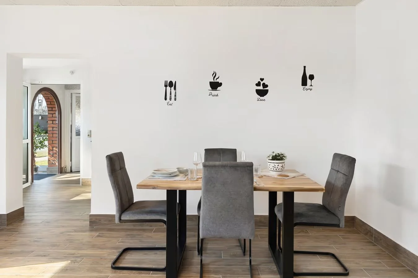 Esszimmer mit vier grauen Stühlen um einen Holztisch, weißer Wand mit schwarzen Dekos mit Worten wie '''Eat''', '''Drink''', '''Love''', '''Enjoy'''; im Hintergrund offener Eingang mit Glas und Bruchsteinbogen, Holzboden.