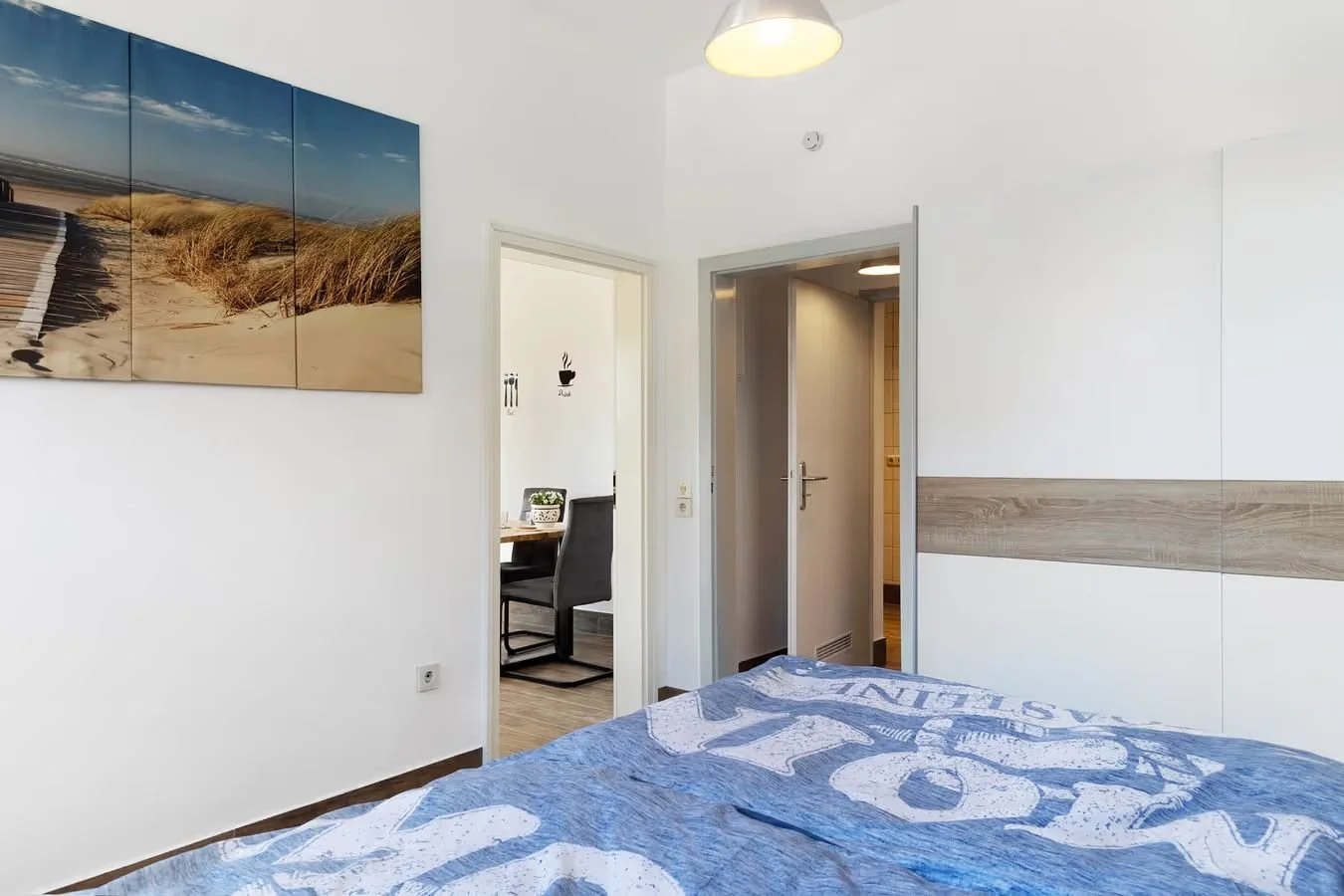 Blick in ein Schlafzimmer mit einem Bett, Wandbilder, und eine Tür zum Badezimmer oder Flur, sowie ein Raum mit einem Tisch und Stühlen im Hintergrund.