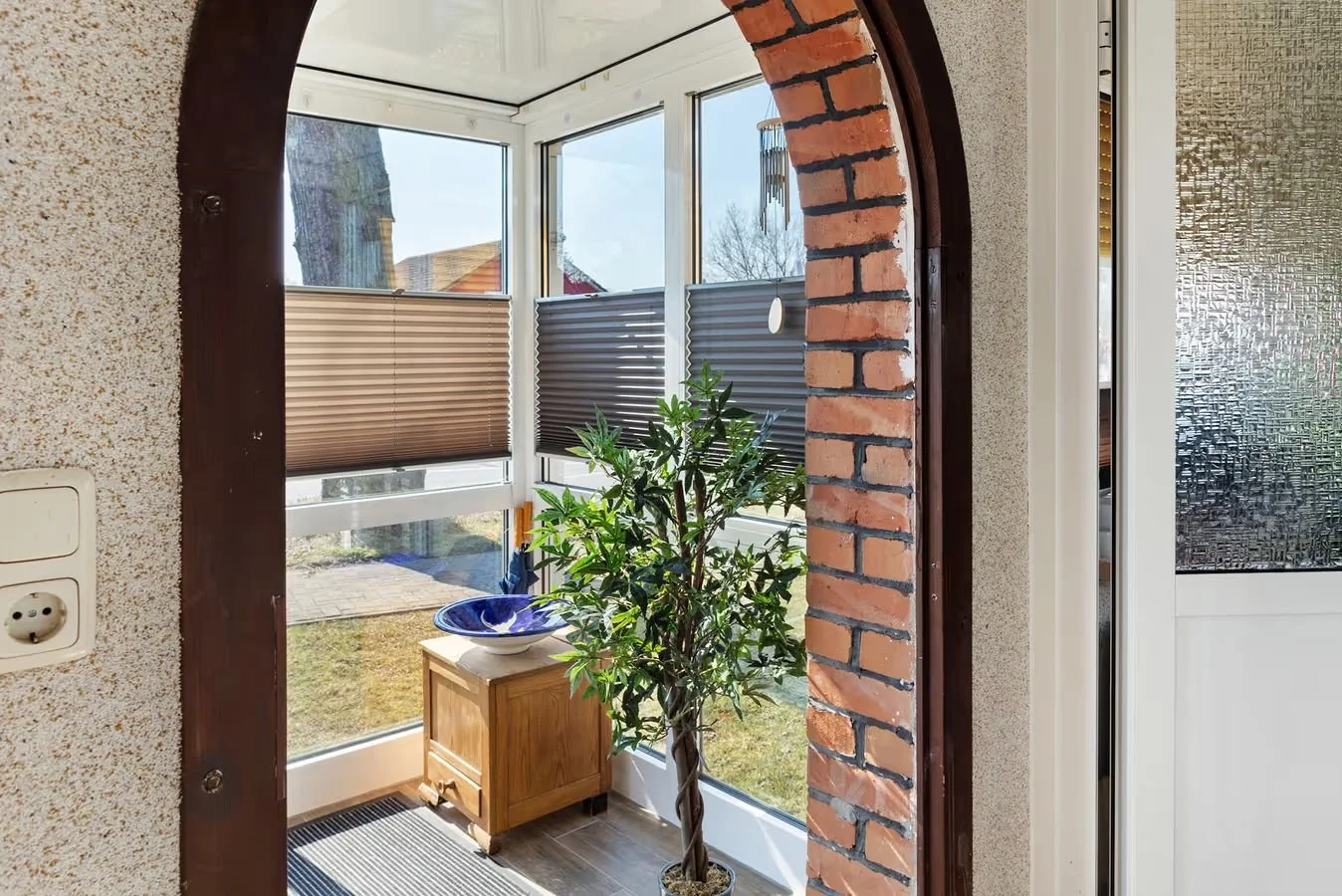 Ein Gewächshaus mit großen Fenstern, einer Zimmerpflanze, einem kleinen Holztisch mit einer blauen Schüssel, Terassenzubehör und einer Gartenansicht draußen.