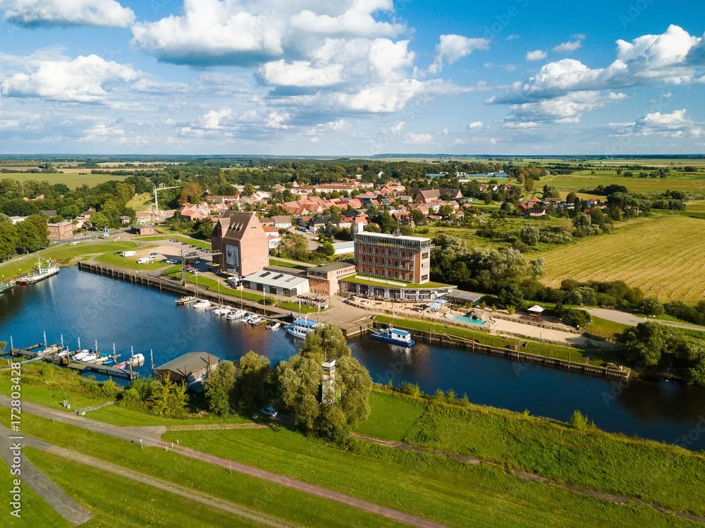 ein Hafen mit mehreren Booten und modernem Gebäude, umgeben von landwirtschaftlichen Feldern und einem kleinen Dorf.
