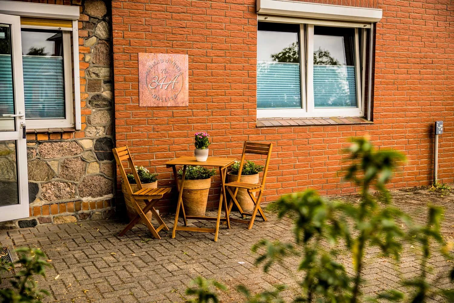 Ein Außenbereich mit einem kleine runden Holztisch und zwei passenden Stühlen. Auf dem Tisch steht eine Blumentopf mit pinkfarbigen Blumen. Es gibt zwei große Tontöpfe mit weißen Blumen neben dem Tisch. Die Wand besteht aus roten Ziegelsteinen und hat ein Schild mit dem Text "Freundliche Wohngemeinschaft in Holli". Ein Fenster mit weißen Rahmen und blauen Fensterläden ist im Hintergrund. Im Vordergrund sind unscharfe grüne Blätter zu sehen.