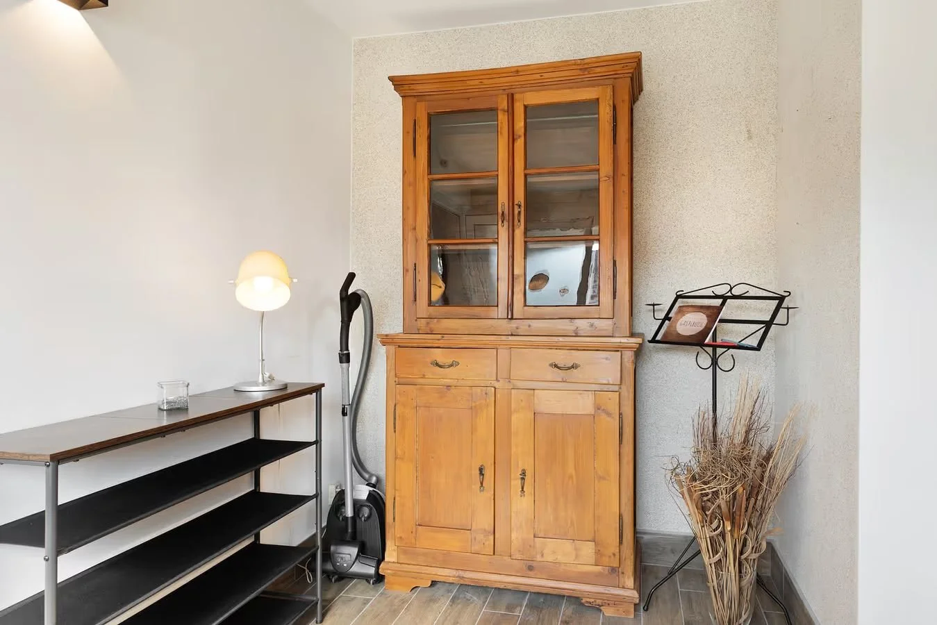 Holzschrank neben einem schwarzen Regal mit einer Lampe, einem Staubsauger, einer Musiknotenständer mit einem Notenblatt, und einem Korb mit trockenen Zweigen.