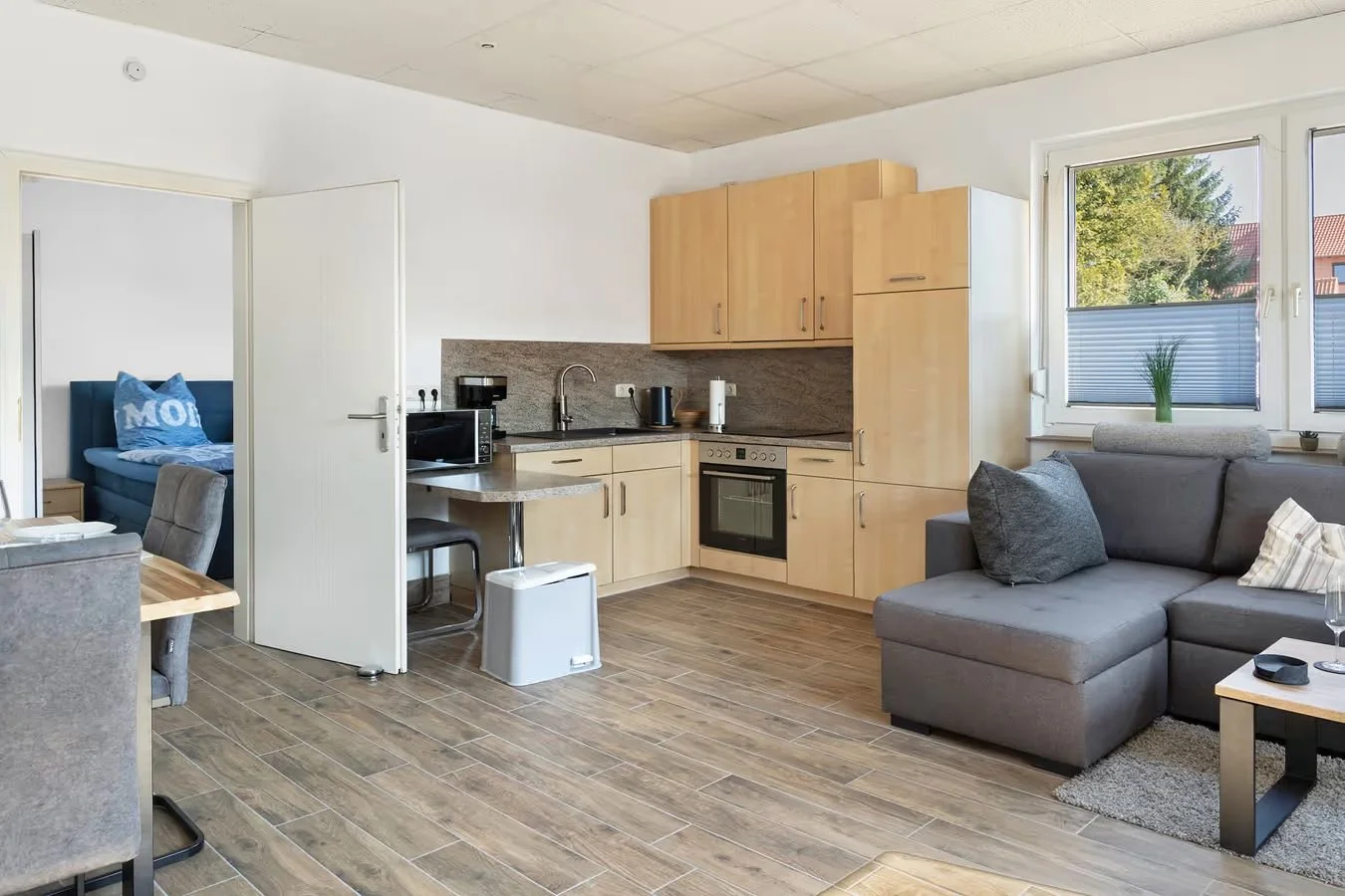 Offene Wohnküche mit Holzküchenschränken, Herd, Mikrowelle, Kühlschrank, Fenster mit Blick auf Bäume, graues Sofa, Kaffeetisch, Wohnzimmer mit Dekorationen.