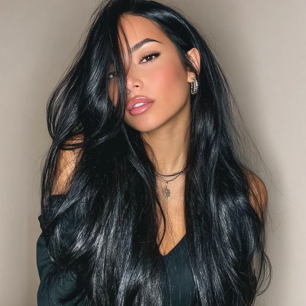 A woman with long, wavy black hair partially covering her face, wearing earrings and a layered necklace, looking directly at the camera with a neutral expression.
