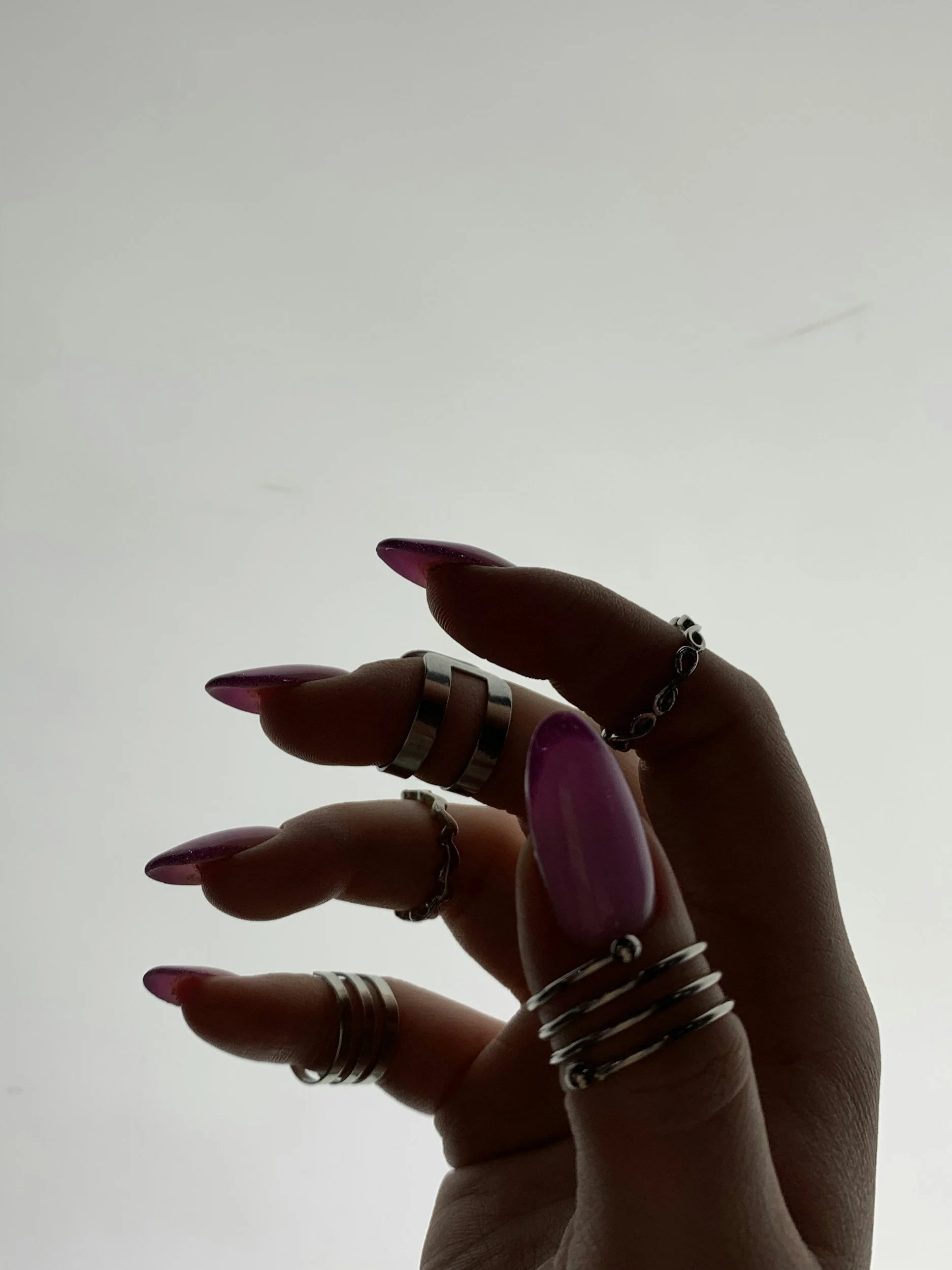 Close-up of a hand with long, painted nails and multiple silver rings, and a delicate chain bracelet, against a plain, light background.