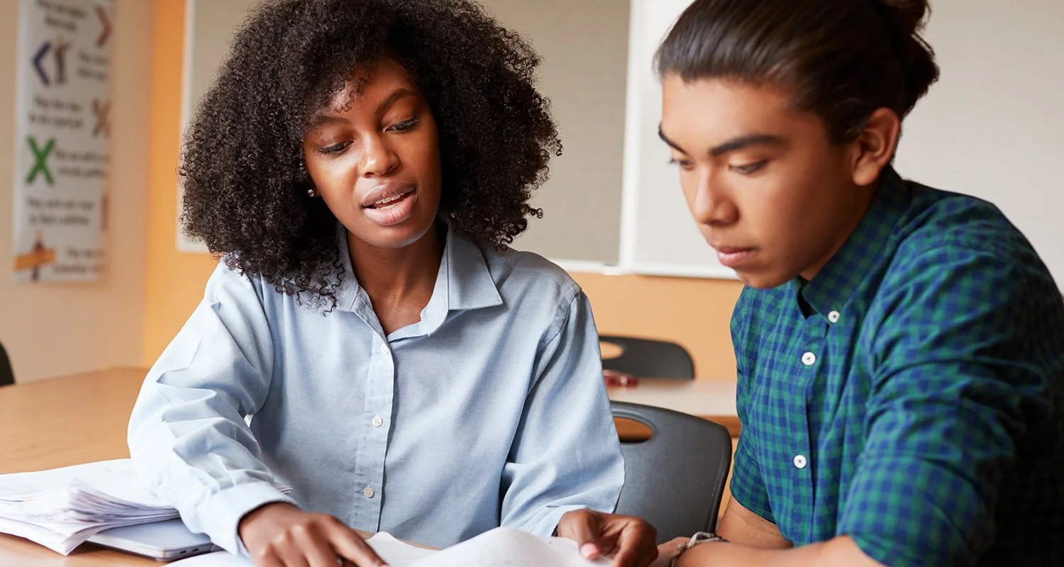 Two students studying together at a school desk, looking at papers.