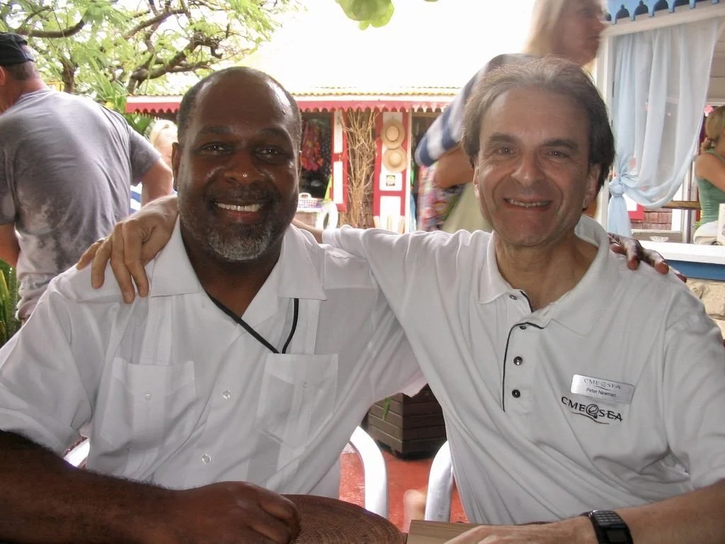 Two men smiling and posing with arms around each other's shoulders at an outdoor gathering, with other people in the background.