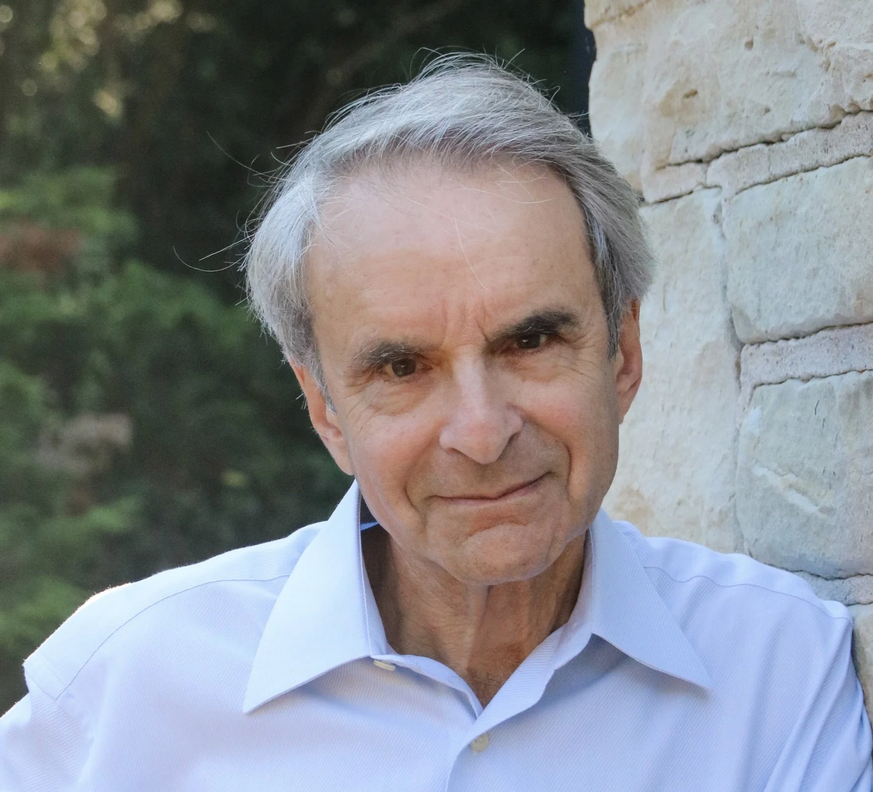 Close-up photo of a smiling man with gray hair, wearing a light blue shirt, outdoors near a brick wall with greenery in the background.