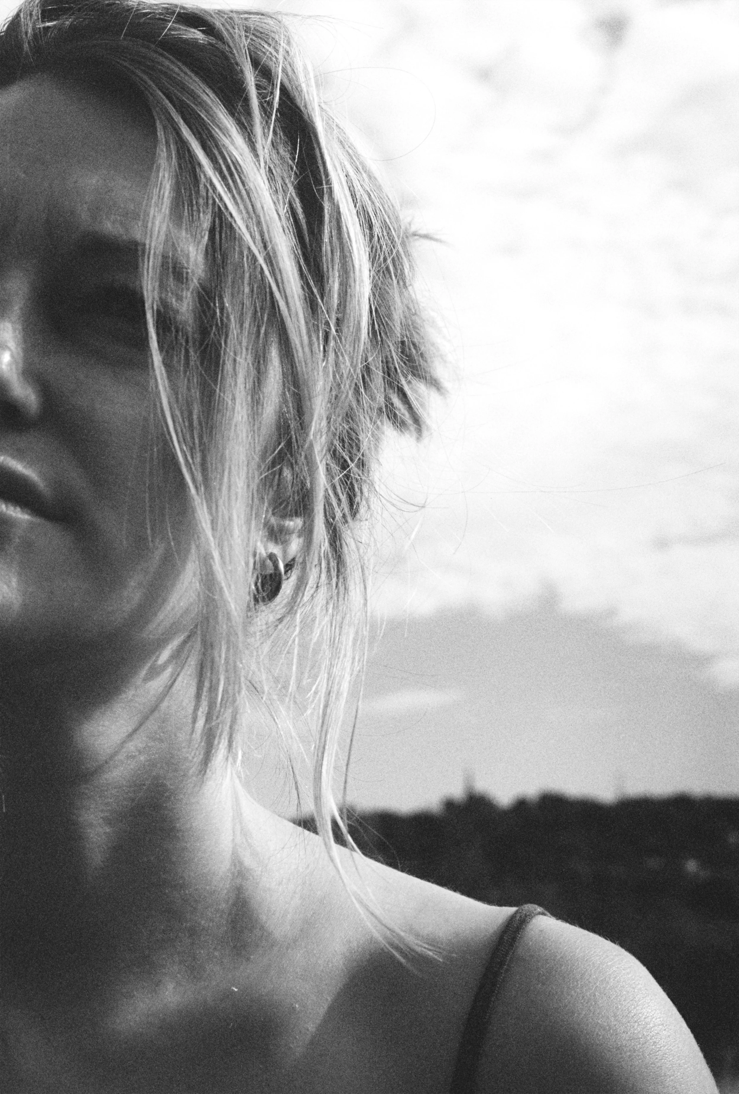 A black-and-white close-up of the left side of a woman's face and shoulder, with her hair partially covering her face. She is wearing a thin strap top or dress, and earrings, with an outdoor background showing a blurry landscape or sky.