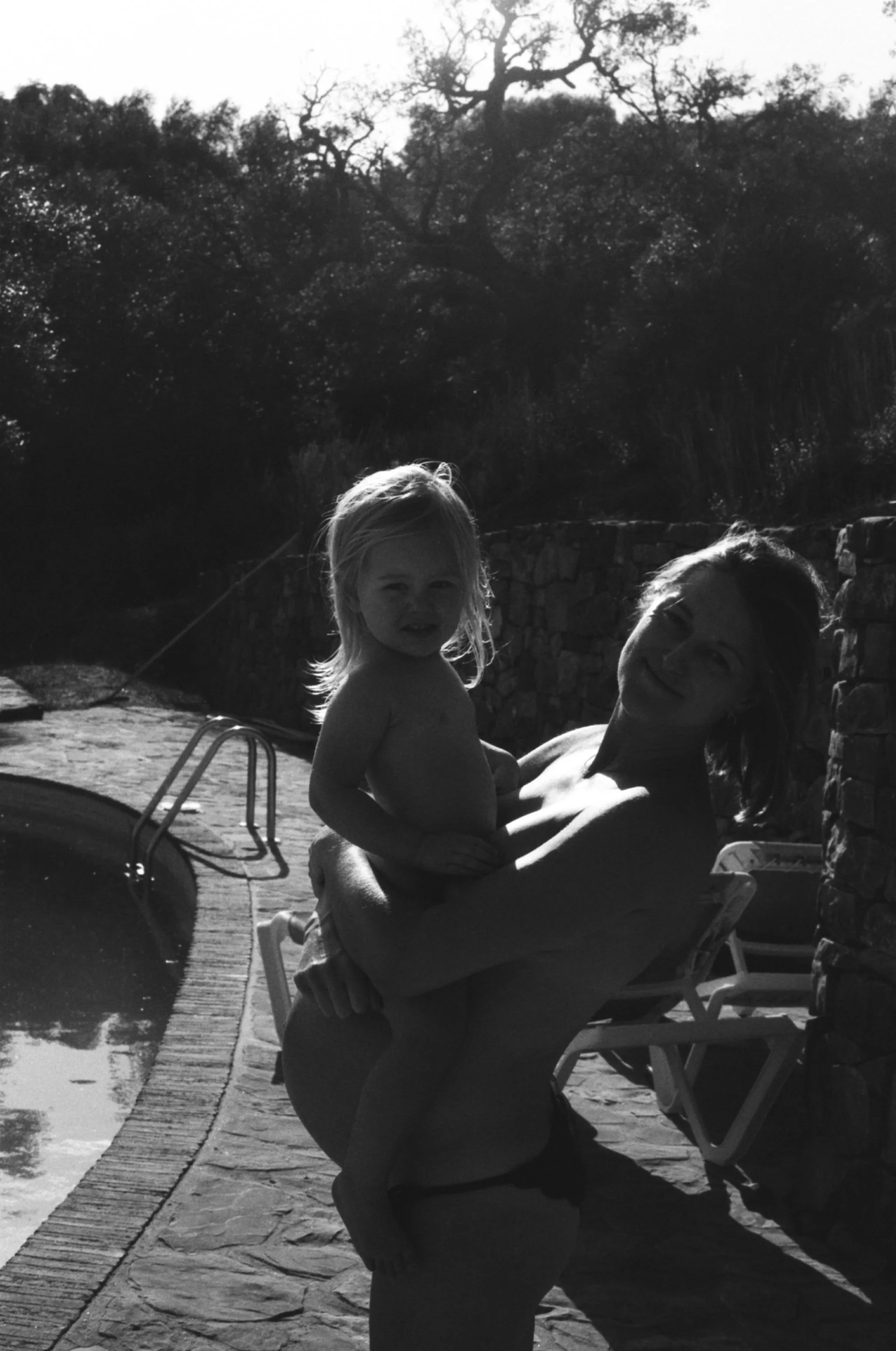 Two people, a woman and a young girl, are outdoors near a swimming pool. The woman is kneeling beside the girl, who is sitting on her lap. The setting includes trees and a stone wall in the background, with the sunlight creating silhouettes and shadows.