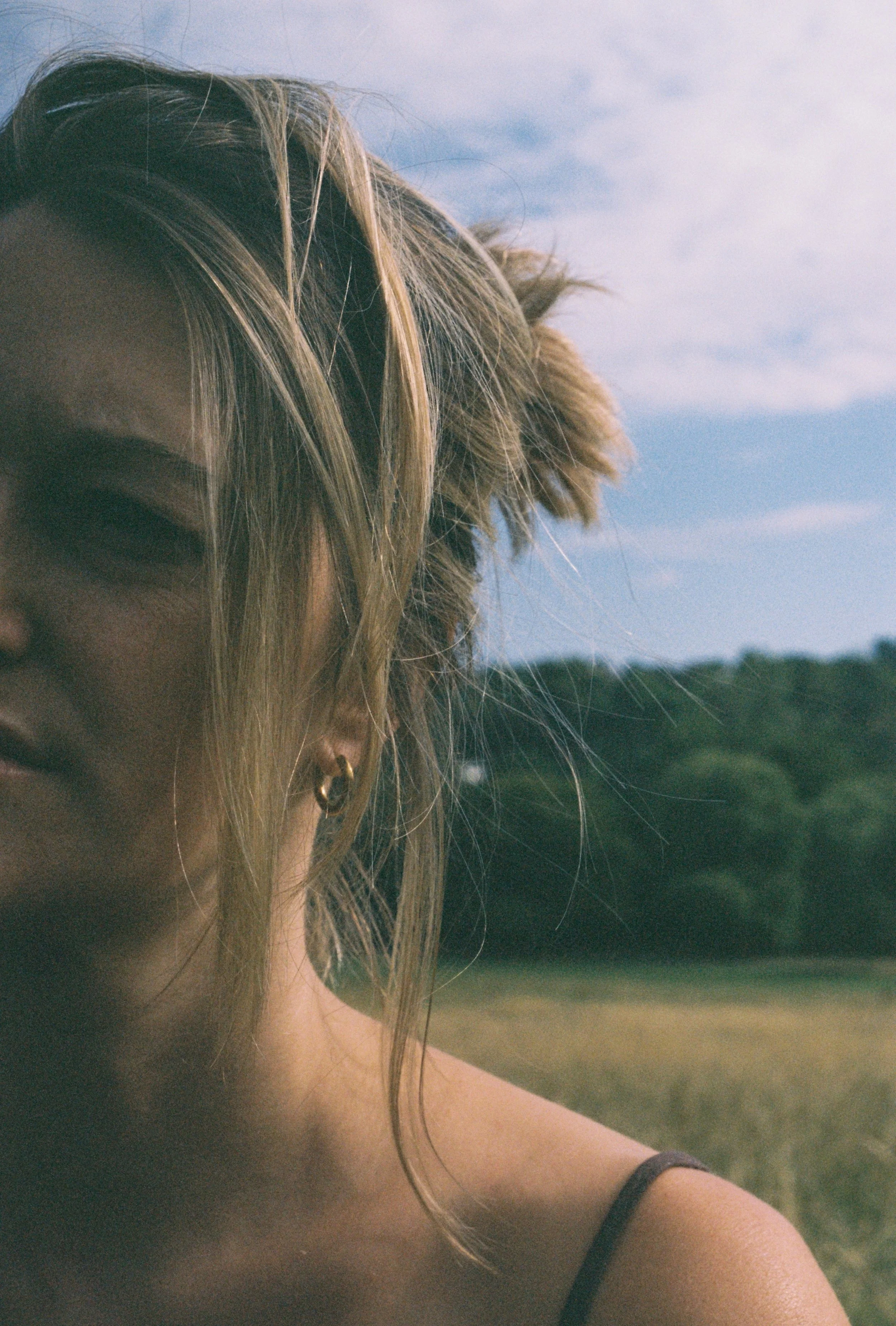 Close-up of a woman's shoulder, hair, and ear with an earring, outdoors with sky and landscape in the background.