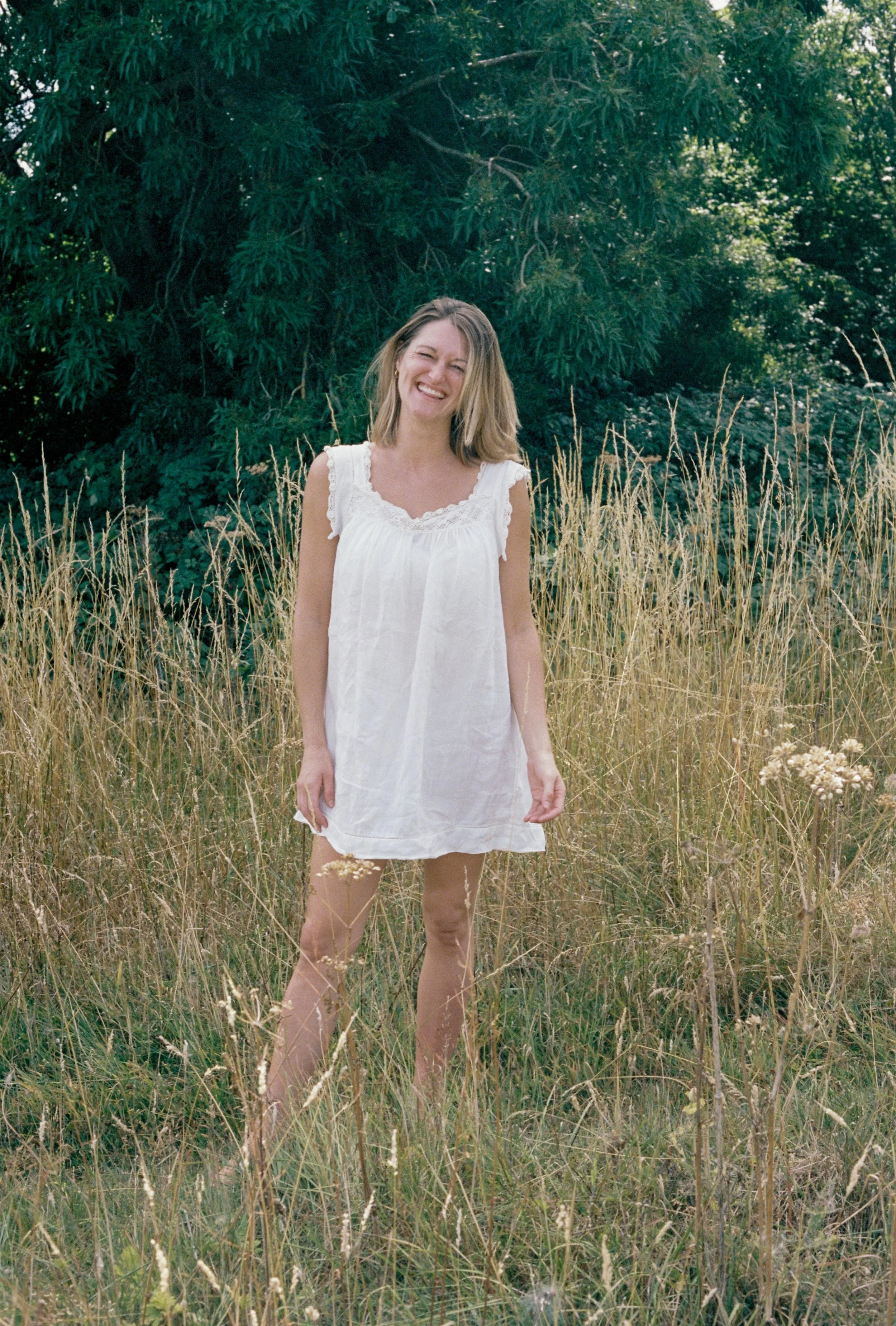 A woman with blonde hair smiling and wearing a white dress standing in a field of tall, dry grass with a large green leafy tree in the background.