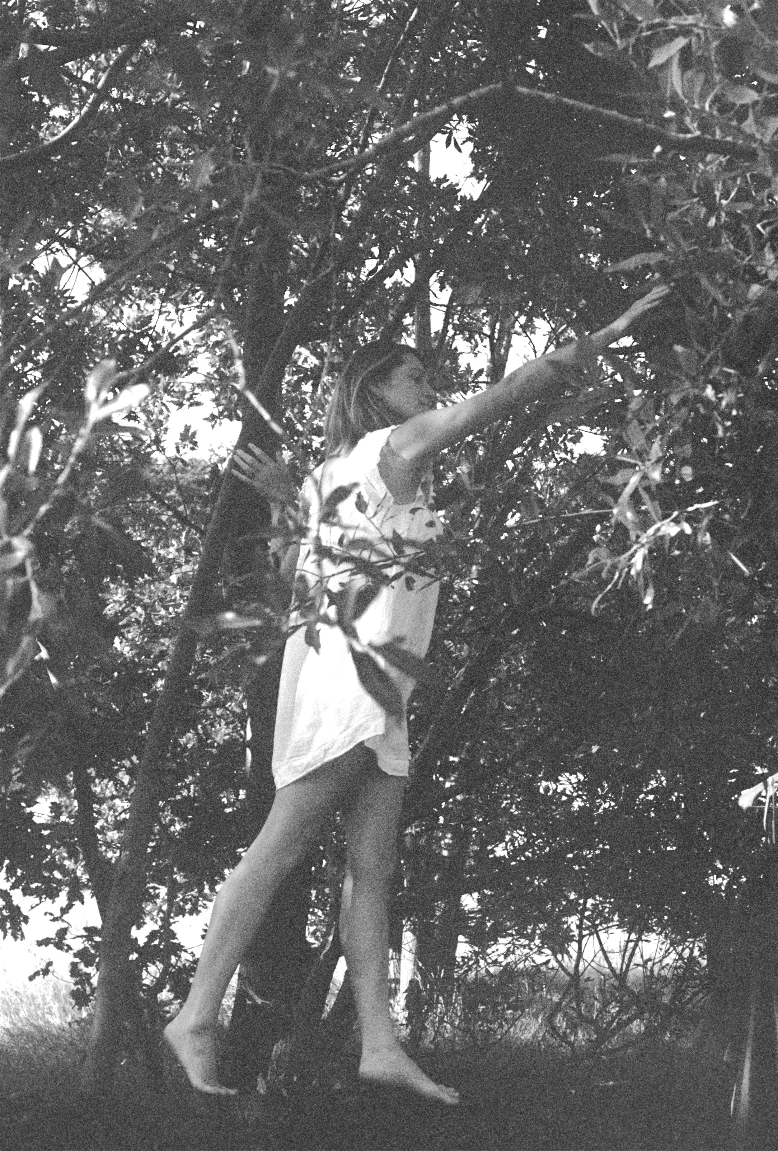 A woman with long hair reaching for leaves or branches on a tree, wearing a light-colored outfit, outdoors in a natural setting with dense foliage.
