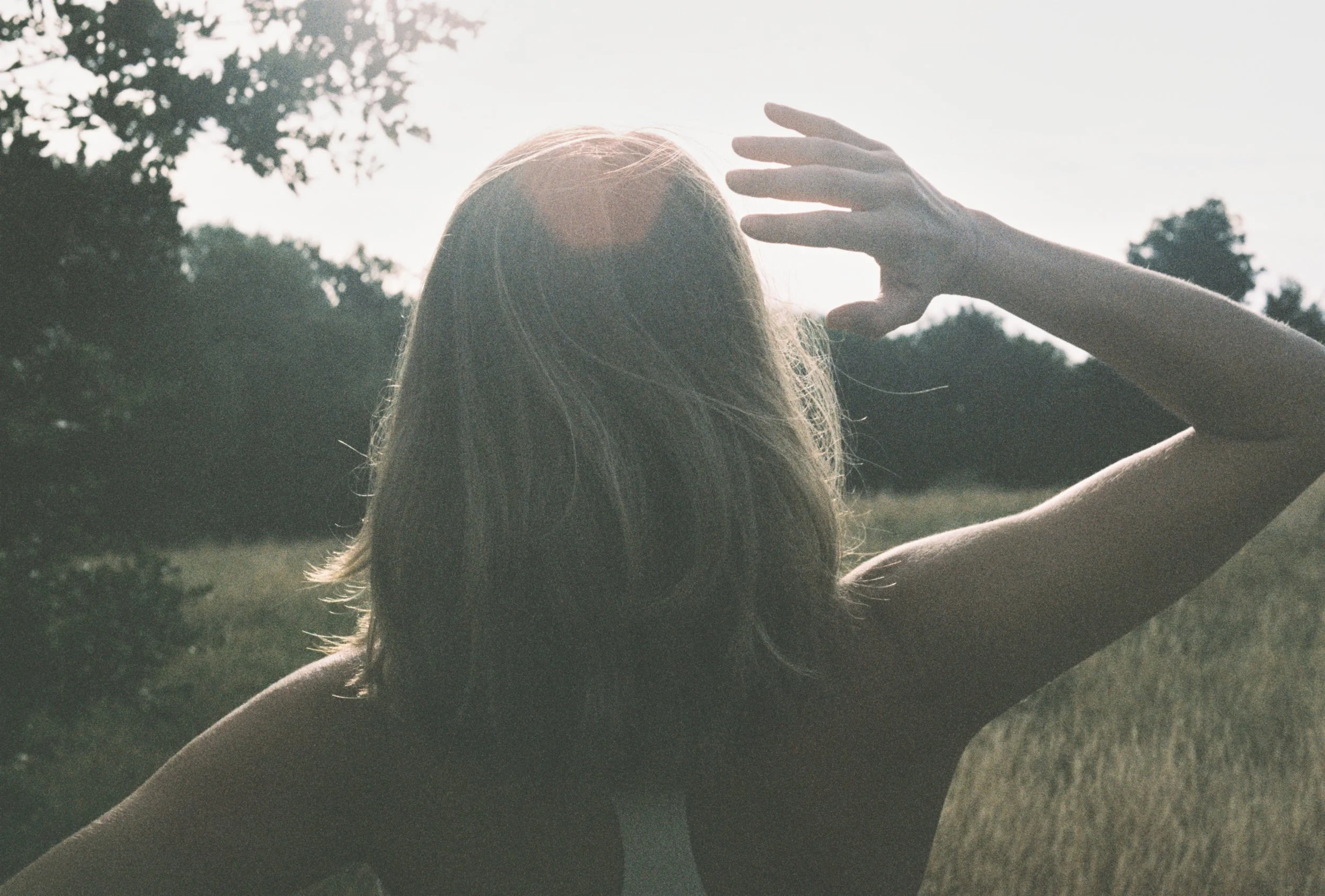 Back view of a woman with long hair outdoors, raising her arm towards the sky during sunset or sunrise with trees in the background.