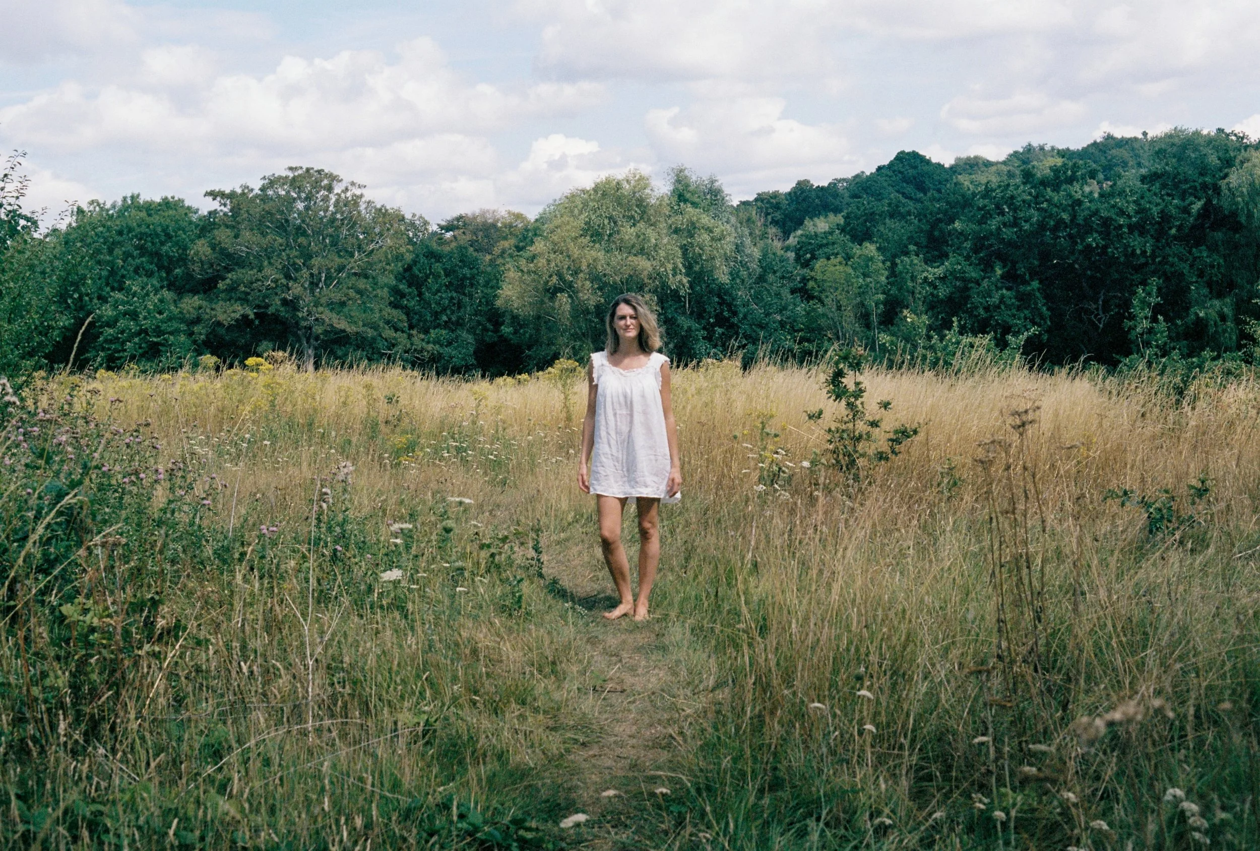A woman in a white dress standing barefoot in a grassy field with trees and cloudy sky in the background.