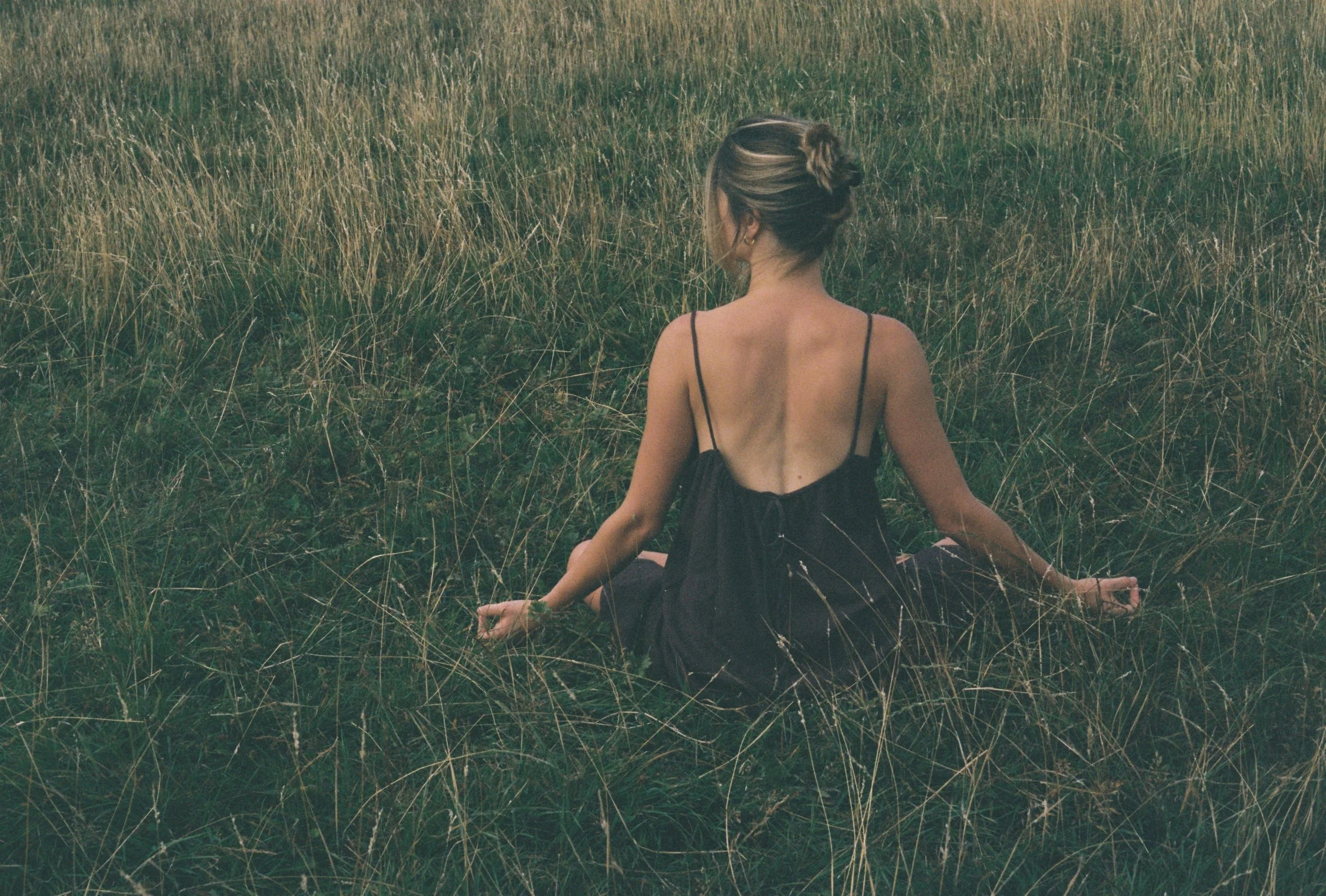 A woman with blonde hair tied in a bun, wearing a black spaghetti strap dress, sitting cross-legged in a grassy field with tall grass.