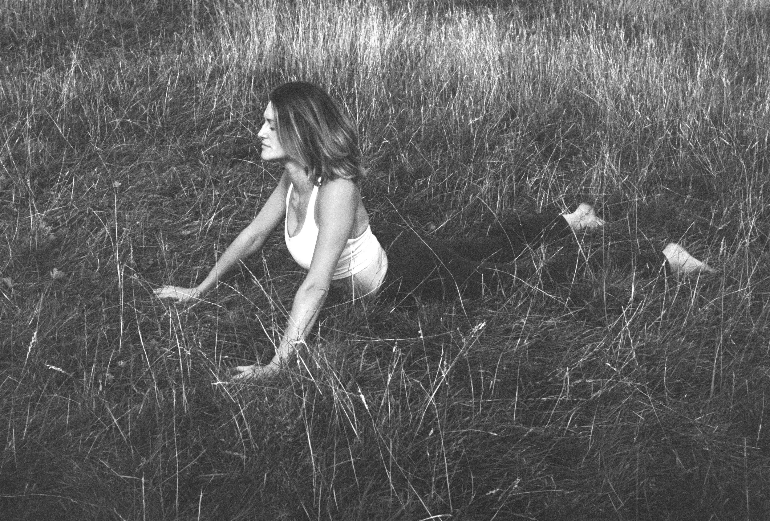 A woman with shoulder-length hair wearing a sleeveless top, sitting on the grass with her legs extended behind her, in a field of tall grass.