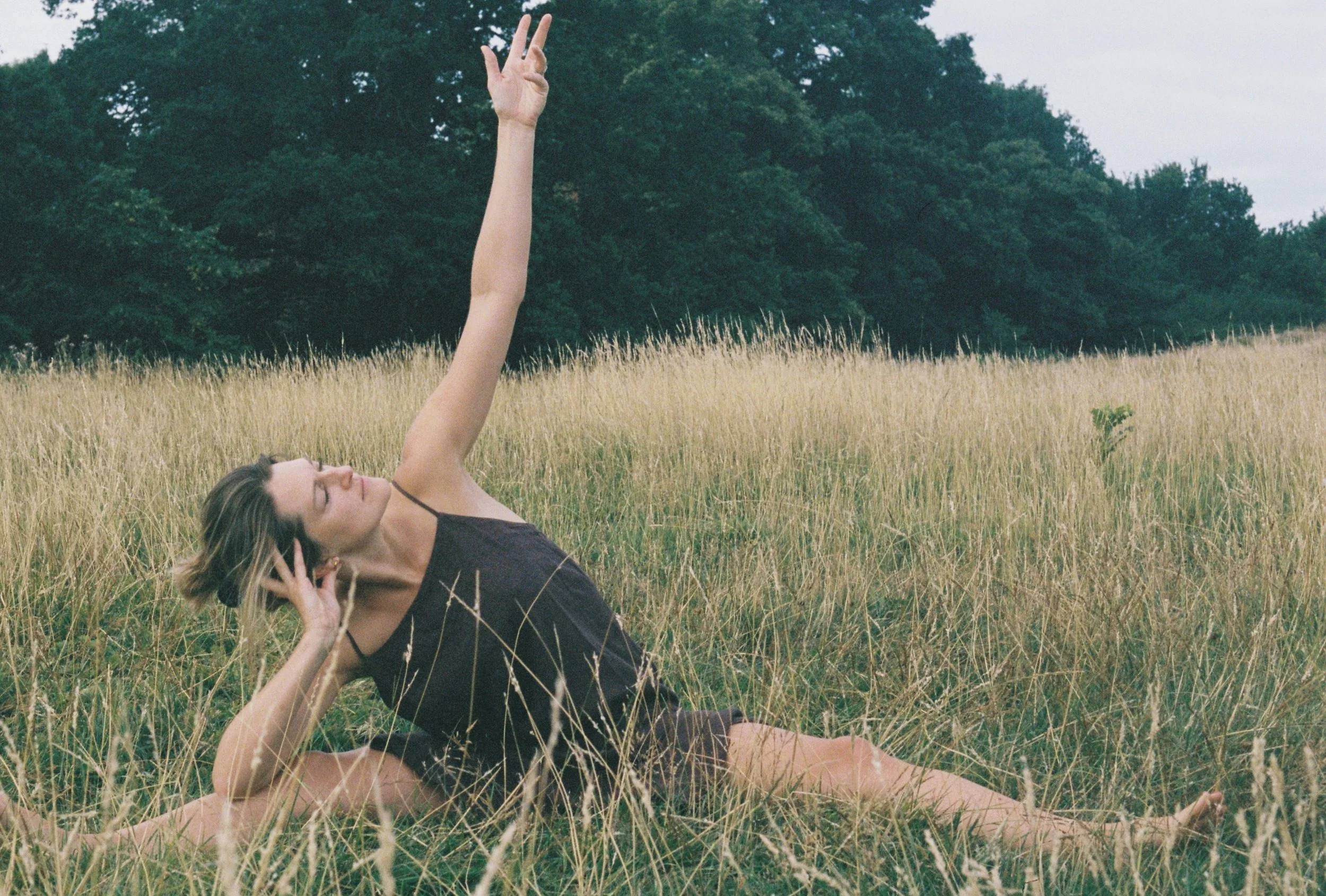 A woman with blonde hair lying in a field of tall grass, with one arm raised and three fingers extended against a background of green trees and cloudy sky.