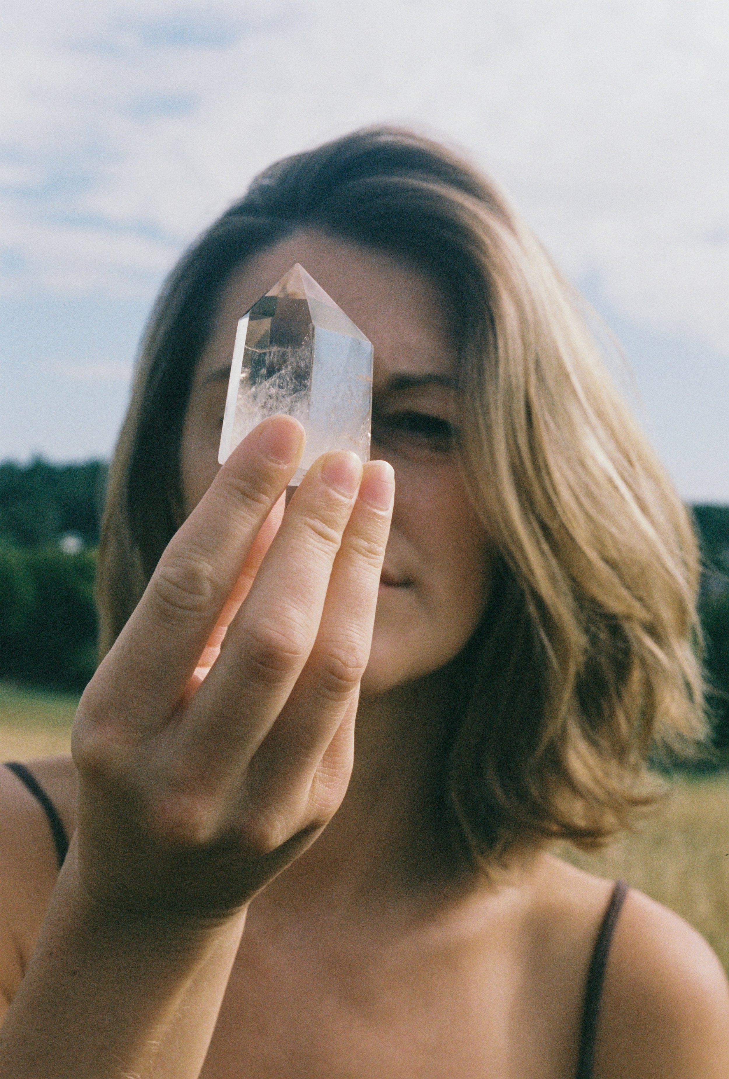 A woman with long, wavy hair holds a clear crystal pointed upwards in front of her face, outdoors against a sky with light clouds.