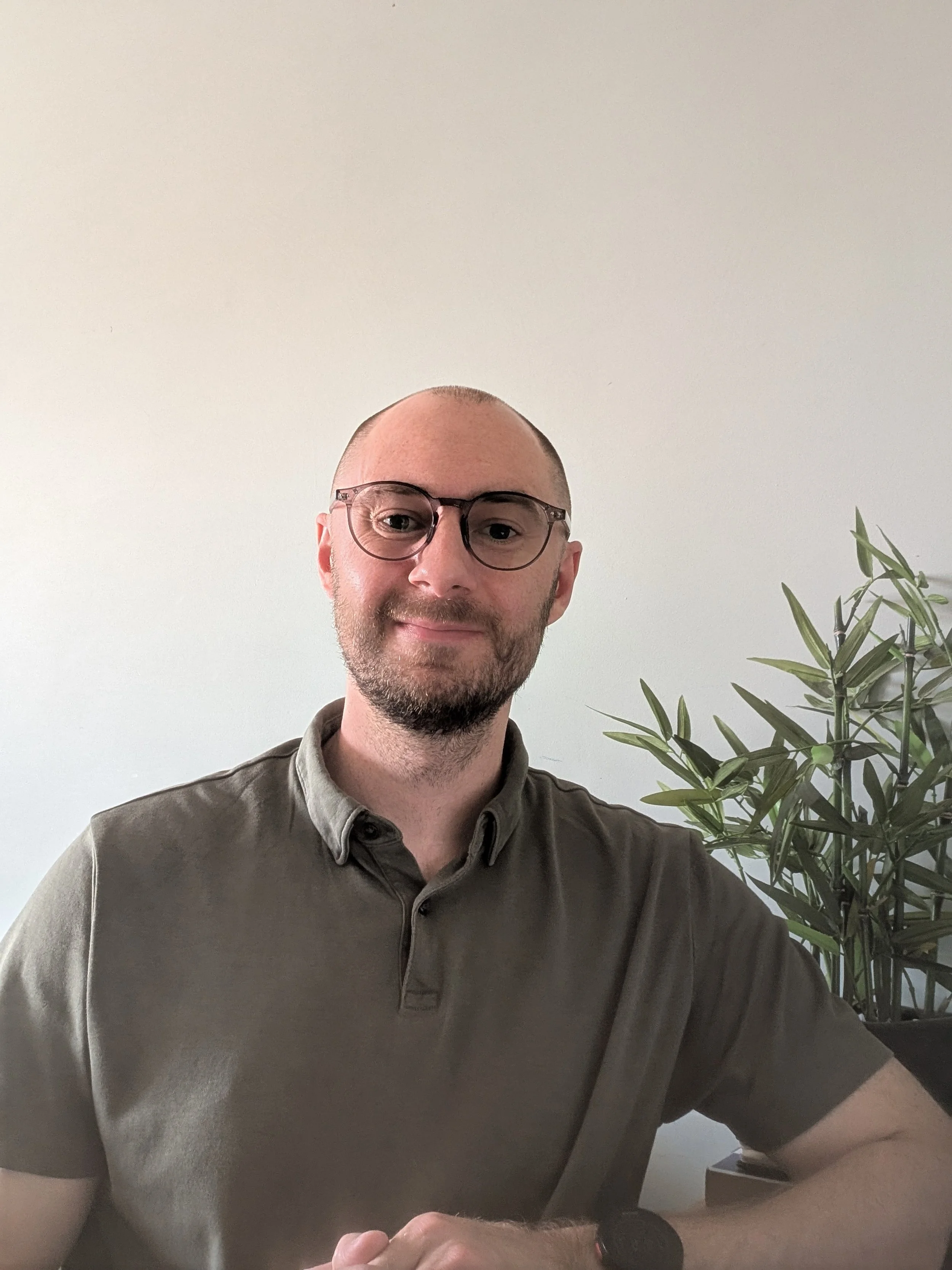 A man with glasses smiling, wearing a dark collared shirt, sitting in front of a plain white wall with a large green plant on the right side.