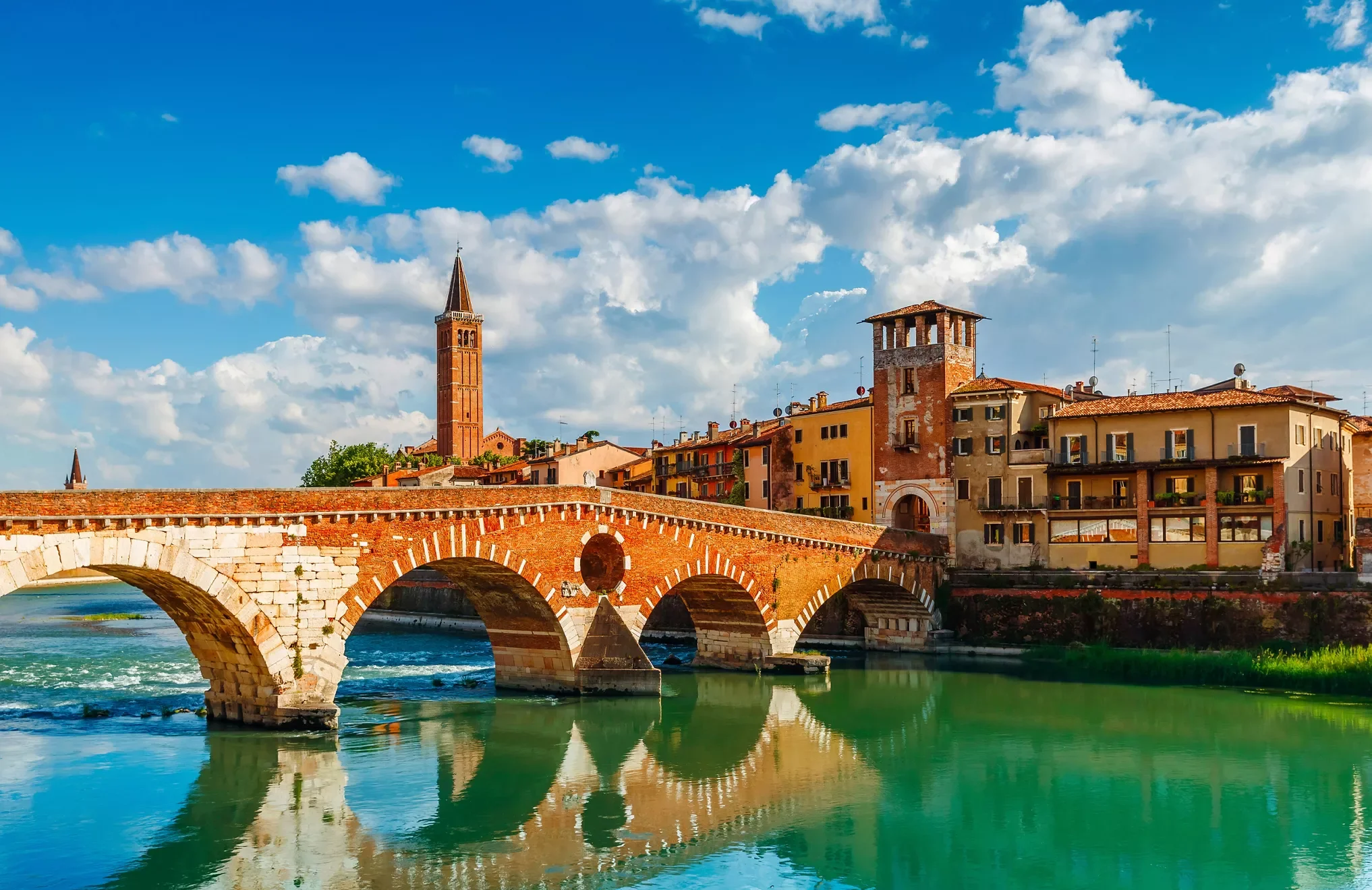 Paesaggio urbano con ponte in mattoni che attraversa un fiume, edificio con torre e campanile di mattoni rossi, cielo con nuvole bianche e azzurre.