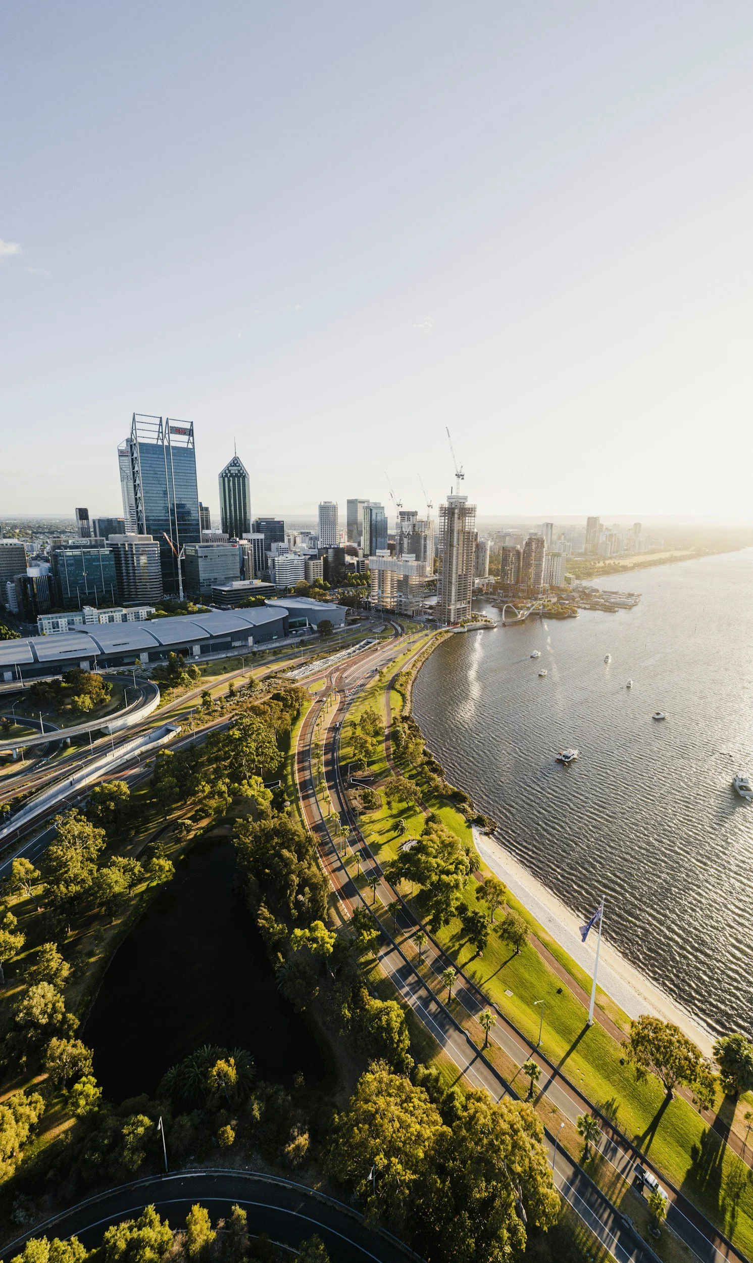 Aerial view of Perth city skyline with skyscrapers, a river with boats, and green parks along the shoreline at sunset.