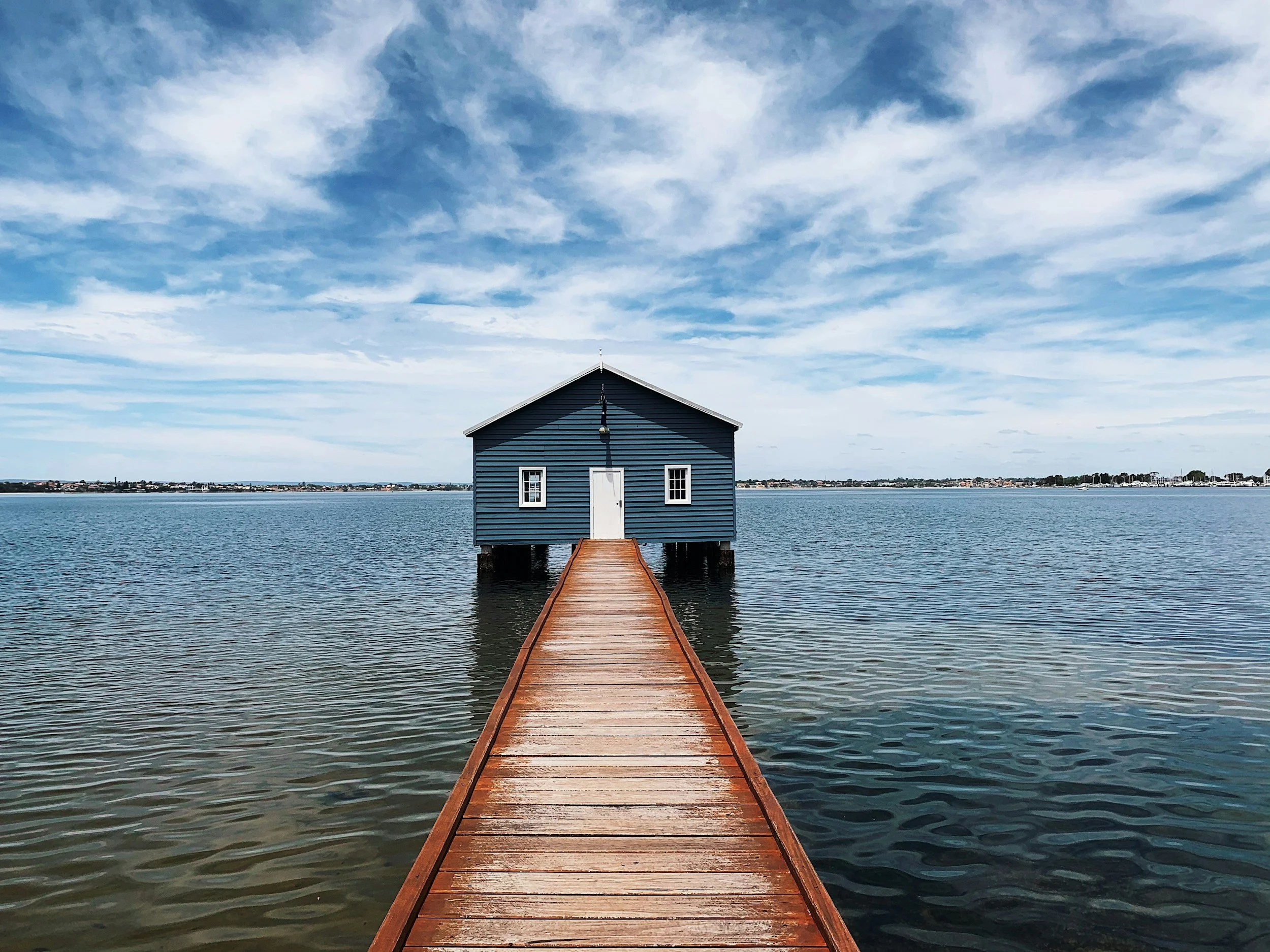 A wooden dock extending over calm water leading to a small blue house with white trim, set against a partly cloudy sky.