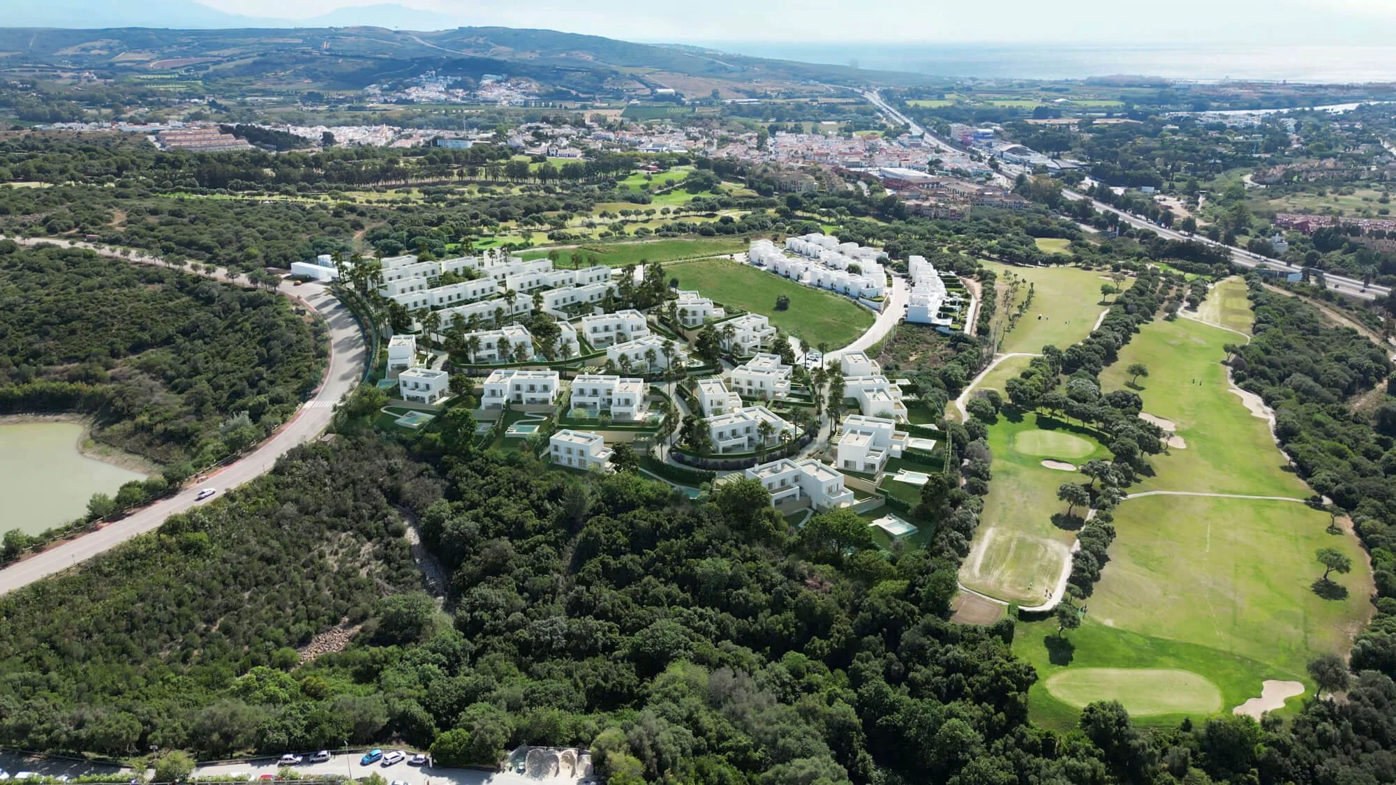 Aerial view of a residential area with white modern houses, a golf course, and lush green trees, surrounded by hills and a city in the background.