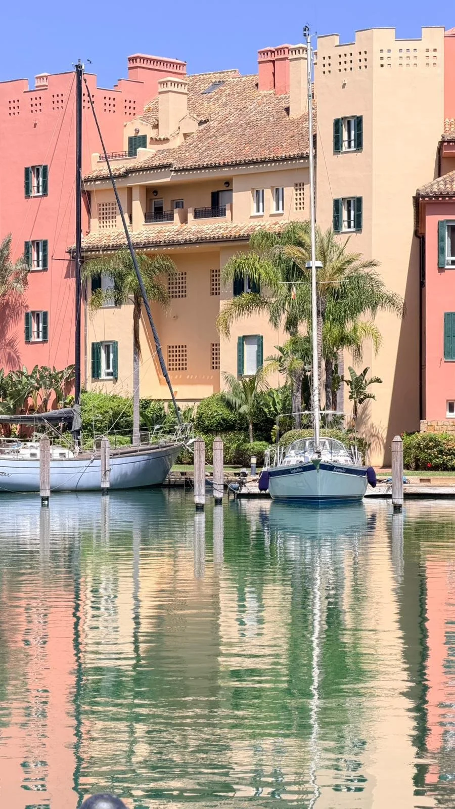 Colorful European-style building with balconies, palm trees, and two sailboats docked in calm water, reflecting the structures.