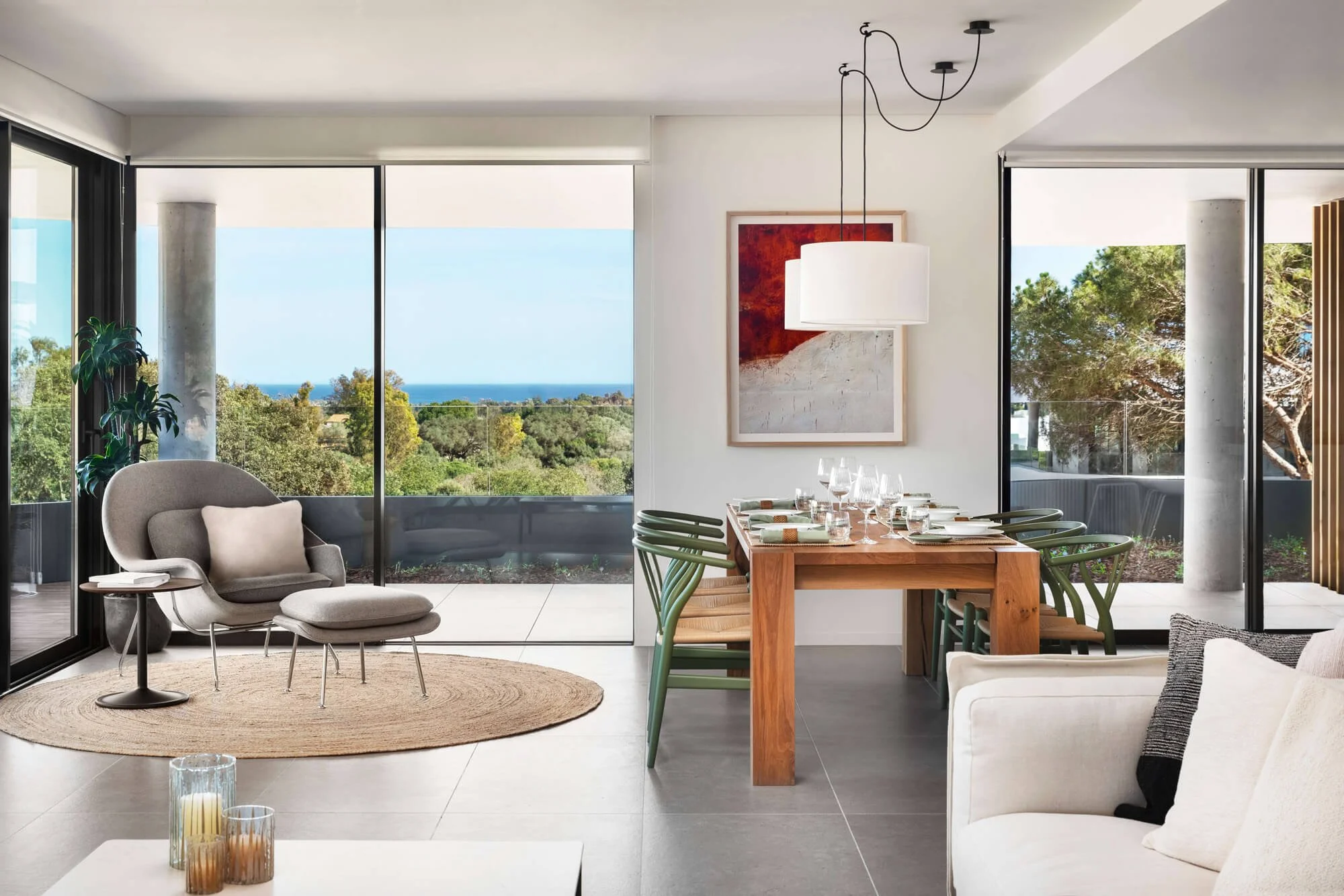 Living and dining area with large windows showing trees and ocean view, modern furniture, artwork, plants, and a neutral color palette.