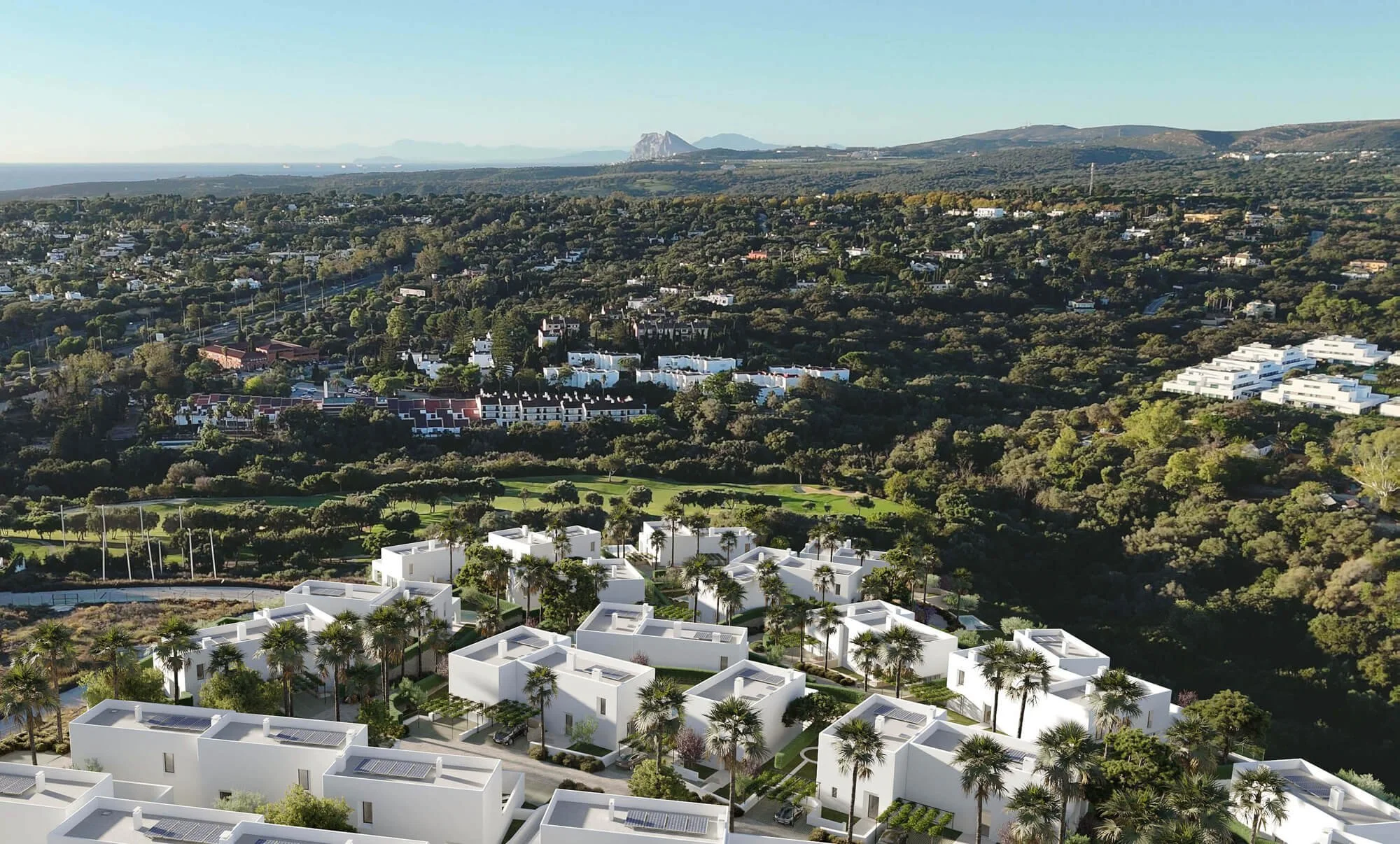 Aerial view of a residential area with white modern houses, green trees, and a golf course in the foreground, with hills and mountains in the background.