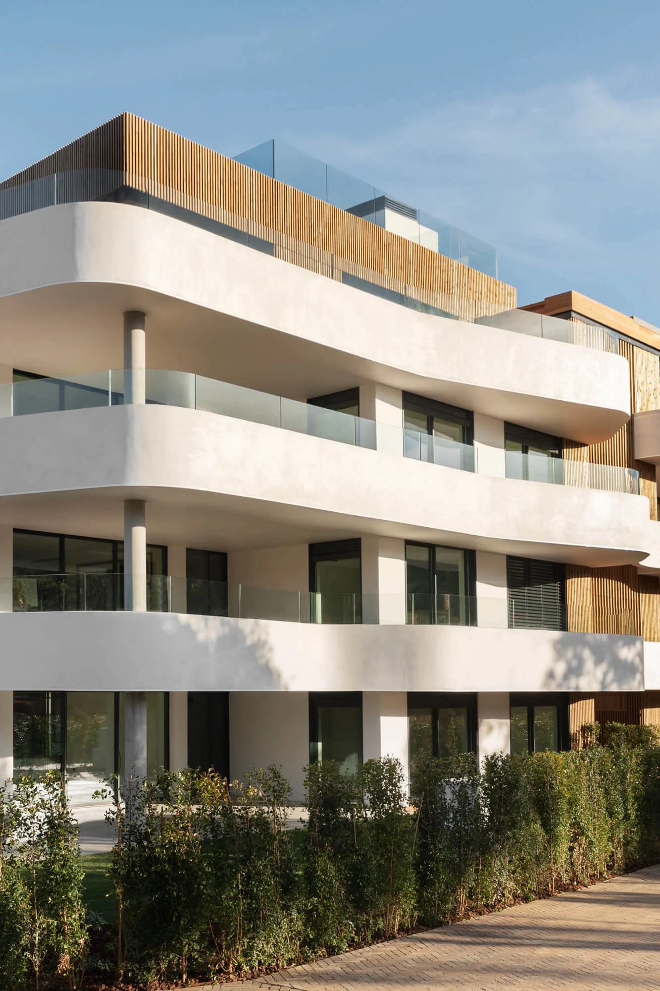Modern multi-story building with white curved balconies, large windows, and wooden accents, surrounded by bushes and a paved sidewalk.