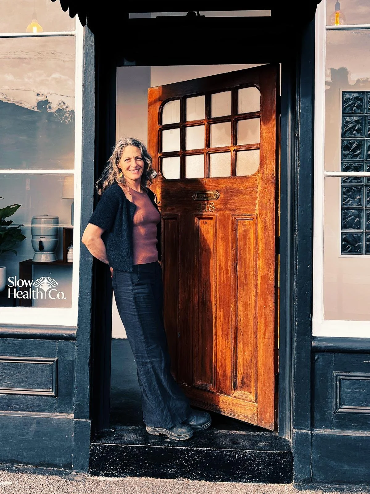 Gina leans out the front of the Well Within in the City space. The front door is an old style wooden door, which is contract with deep blue framing. Gina is wearing a deep blue outfit.